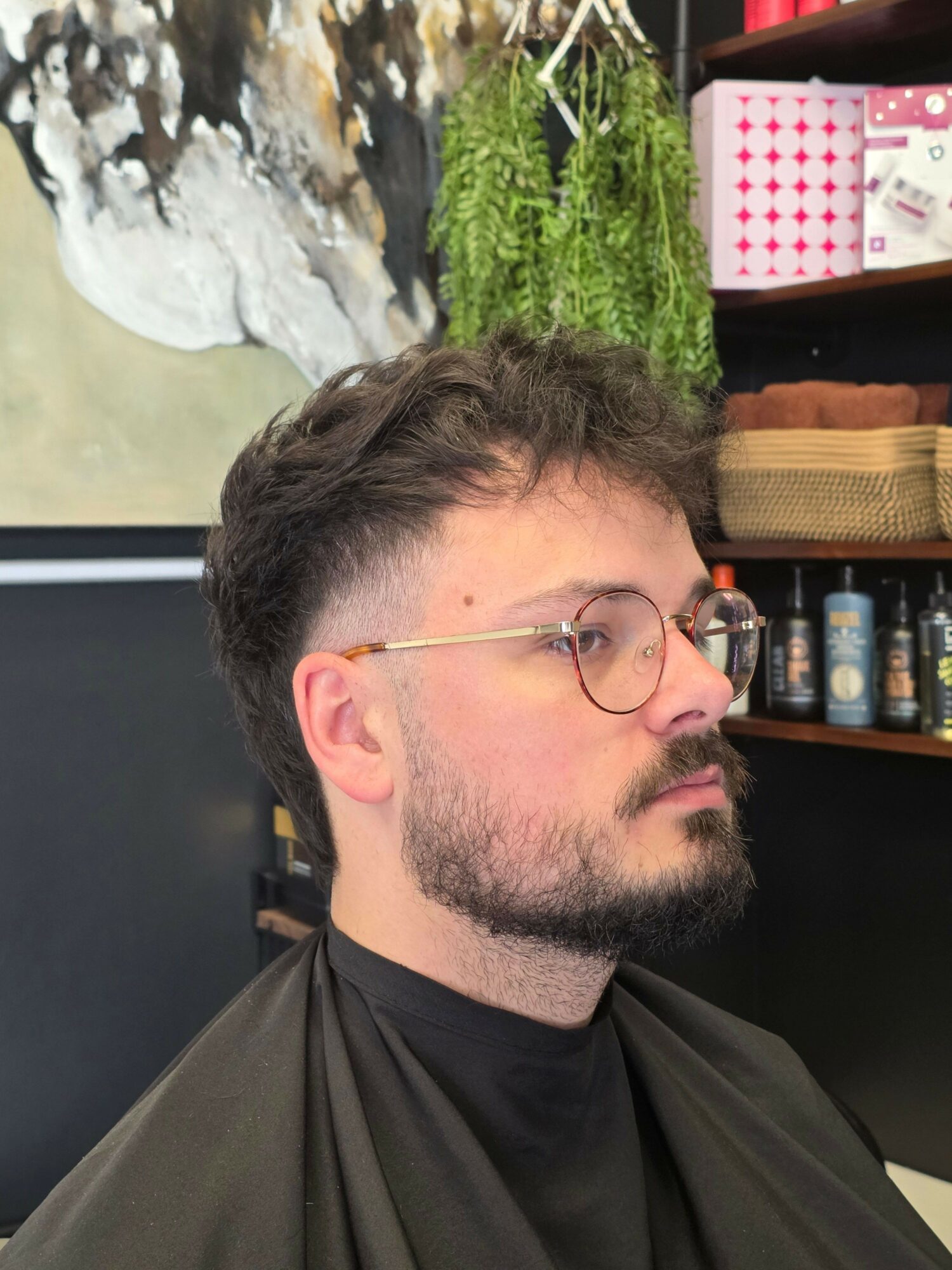 Side view of a man with glasses, beard, and dark hair, sitting outdoors near a waterfall and greenery.