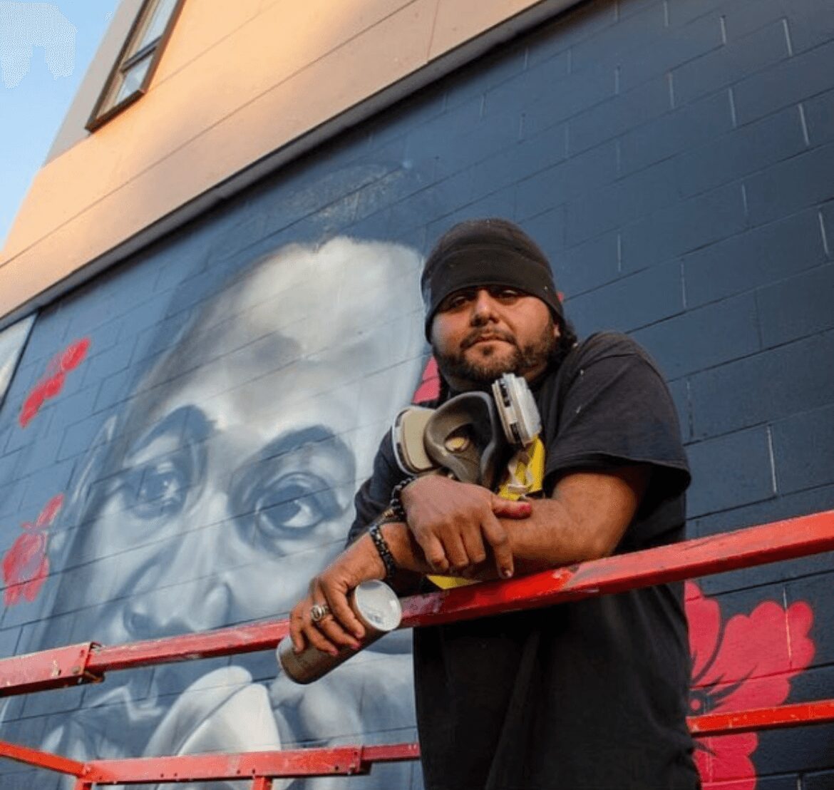Man with a beard and black beanie leaning on a red railing in front of a mural of a person's face on a blue wall.