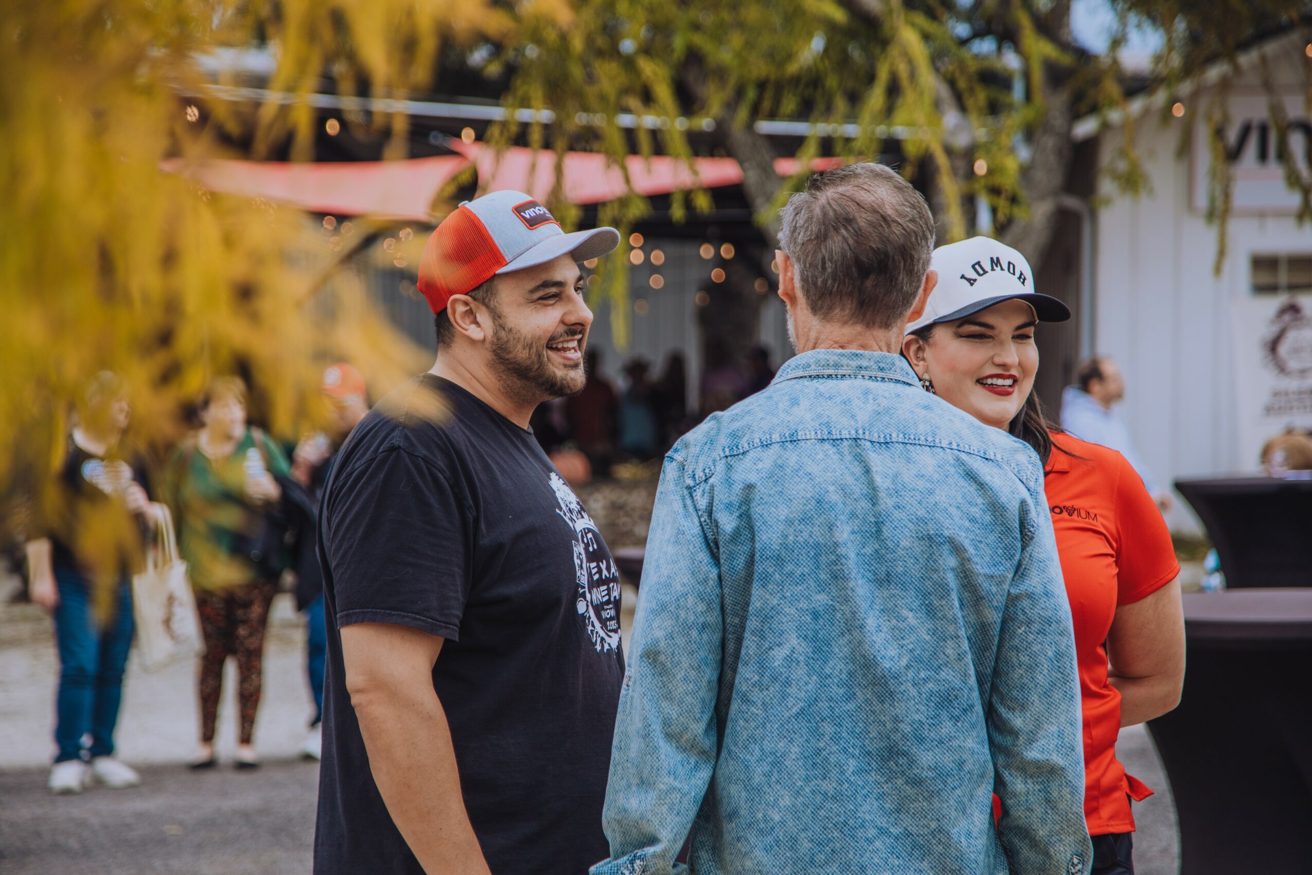 Three people are talking outdoors, with trees and other people in the background, smiling and engaging with each other.