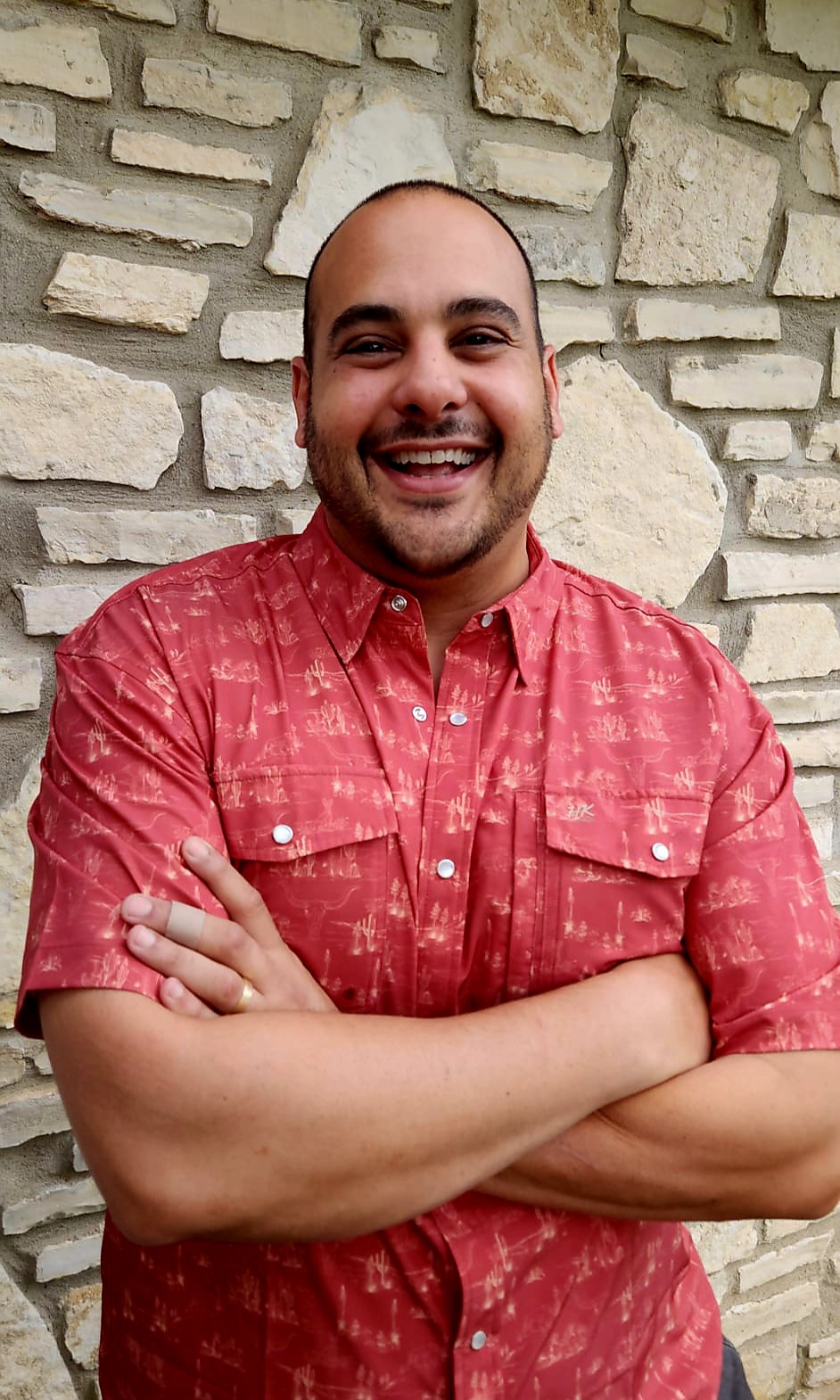 Smiling man with crossed arms wearing a red patterned shirt in front of a stone wall.