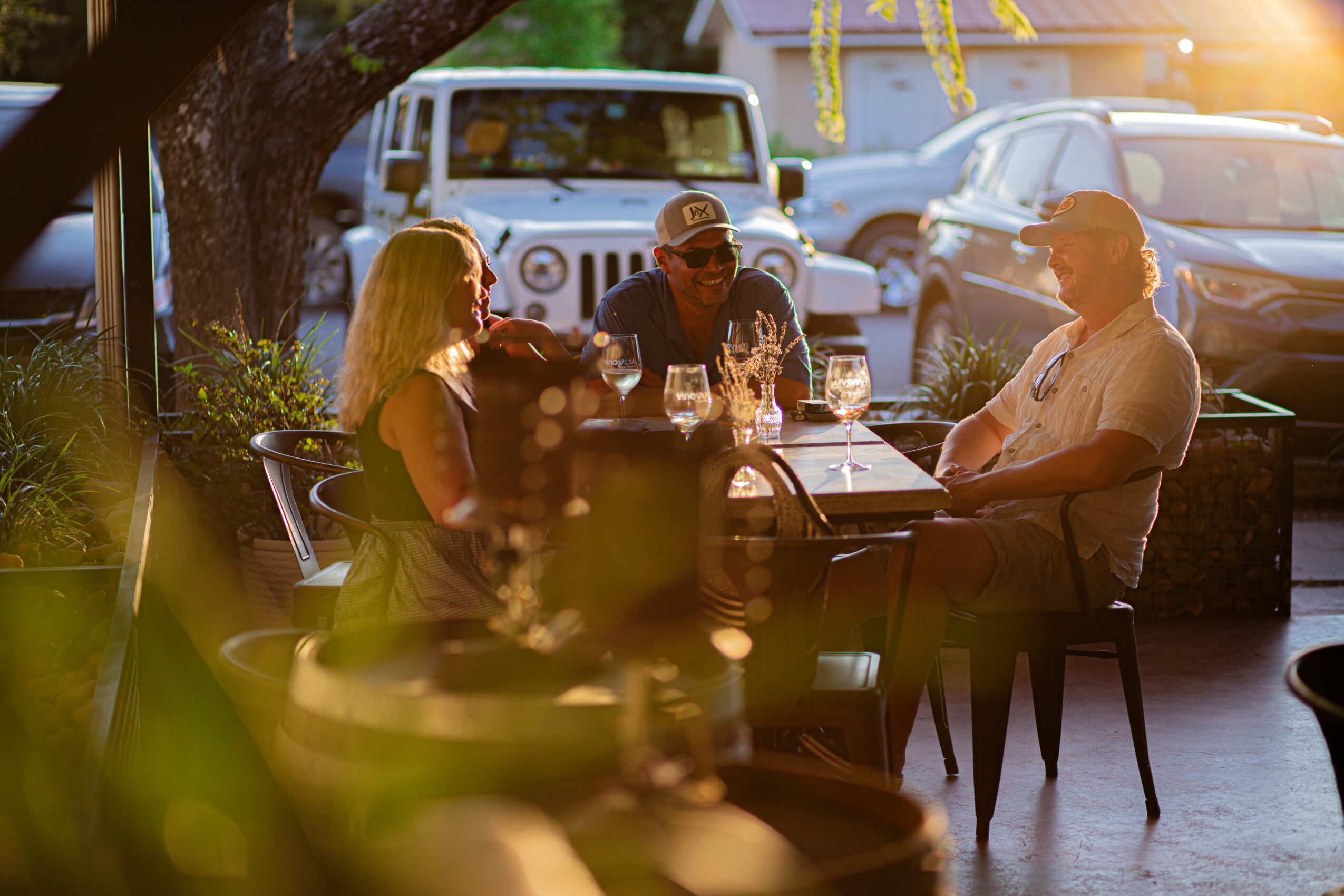 Four people sit at a table outdoors, with cars and a tree in the background, sunlight shining through.