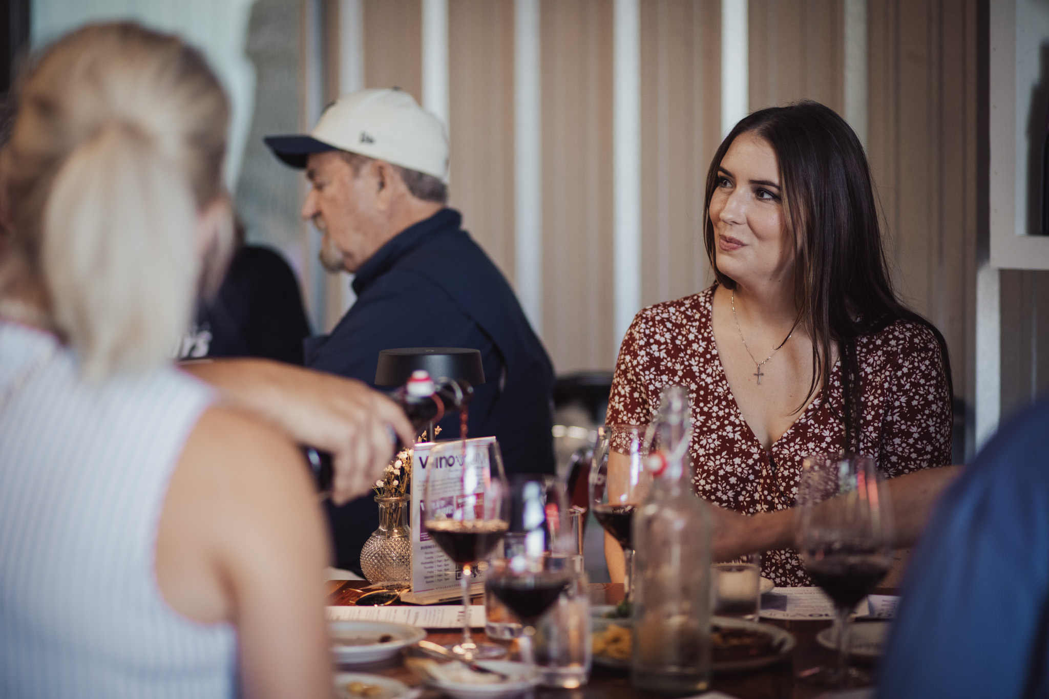 Group of people sitting at a table with drinks, woman with long dark hair smiling, others blurred, indoor setting.