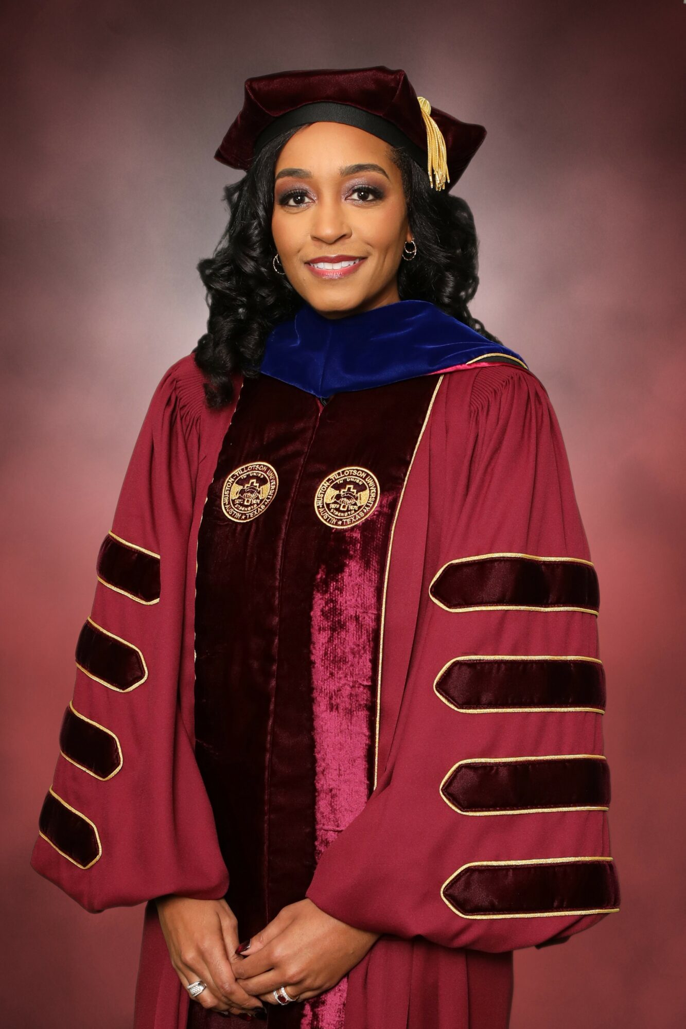 Woman in academic regalia with a maroon and gold gown and a matching cap, smiling, against a pinkish background.