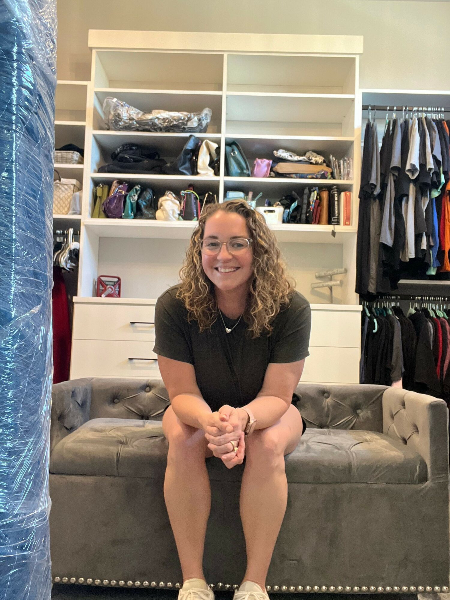Woman sitting on a gray sofa in a walk-in closet with shelves of clothes behind her.