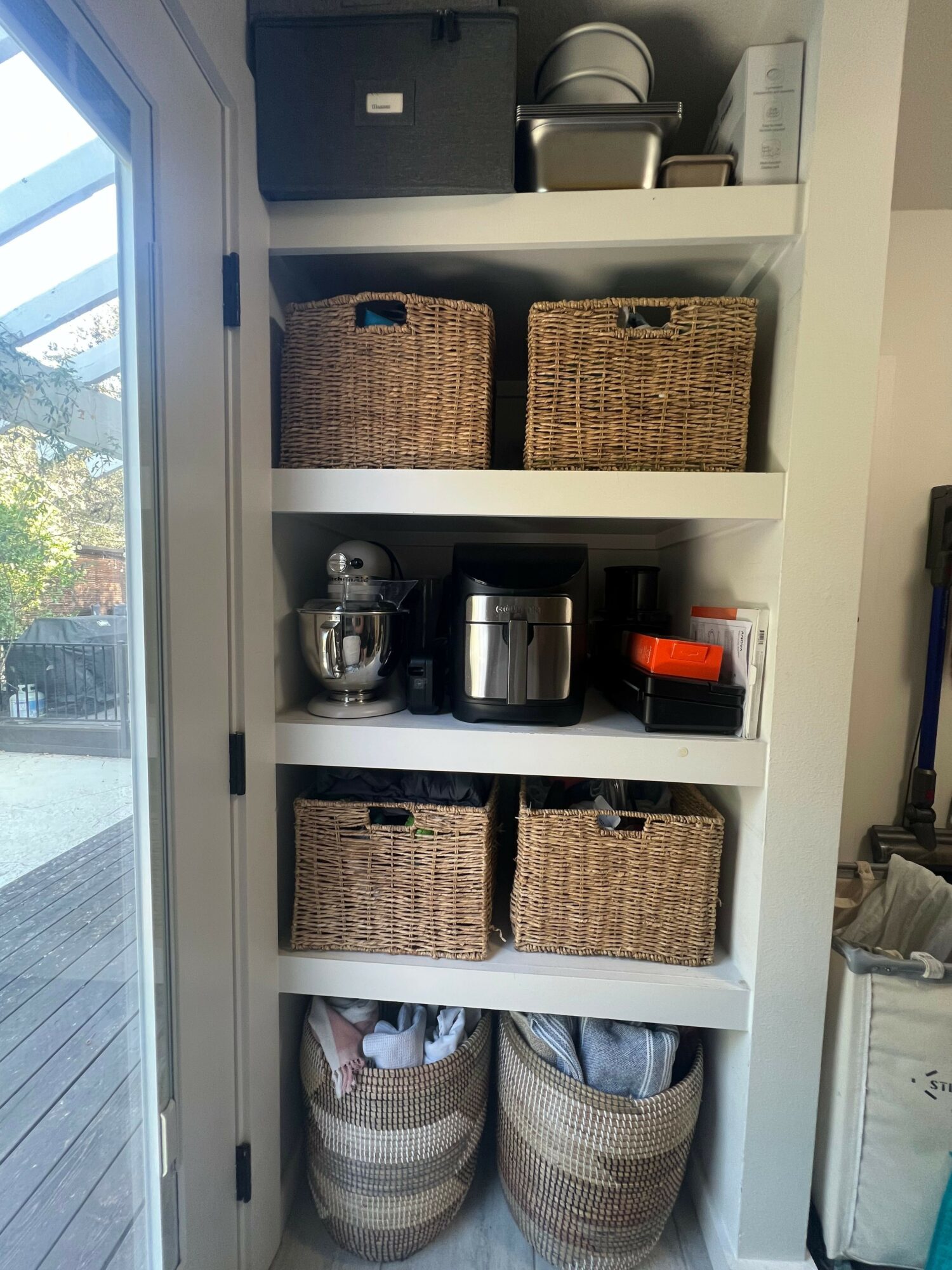 White shelving unit with woven baskets, kitchen appliances, and rolled towels, positioned vertically near a window.