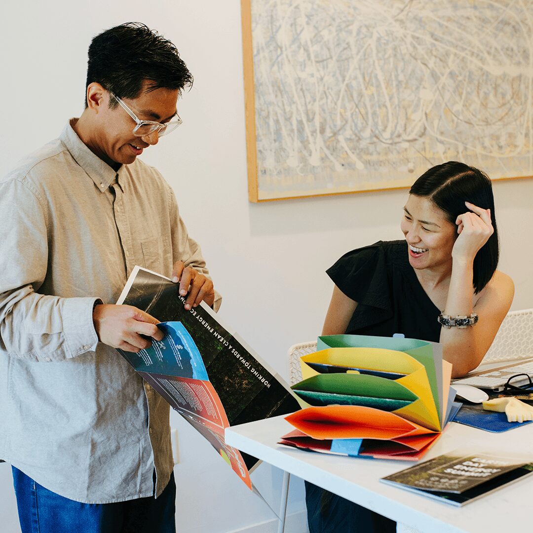 Two people smiling and talking in an office, one holding a magazine, the other sitting at a desk with colorful folders.