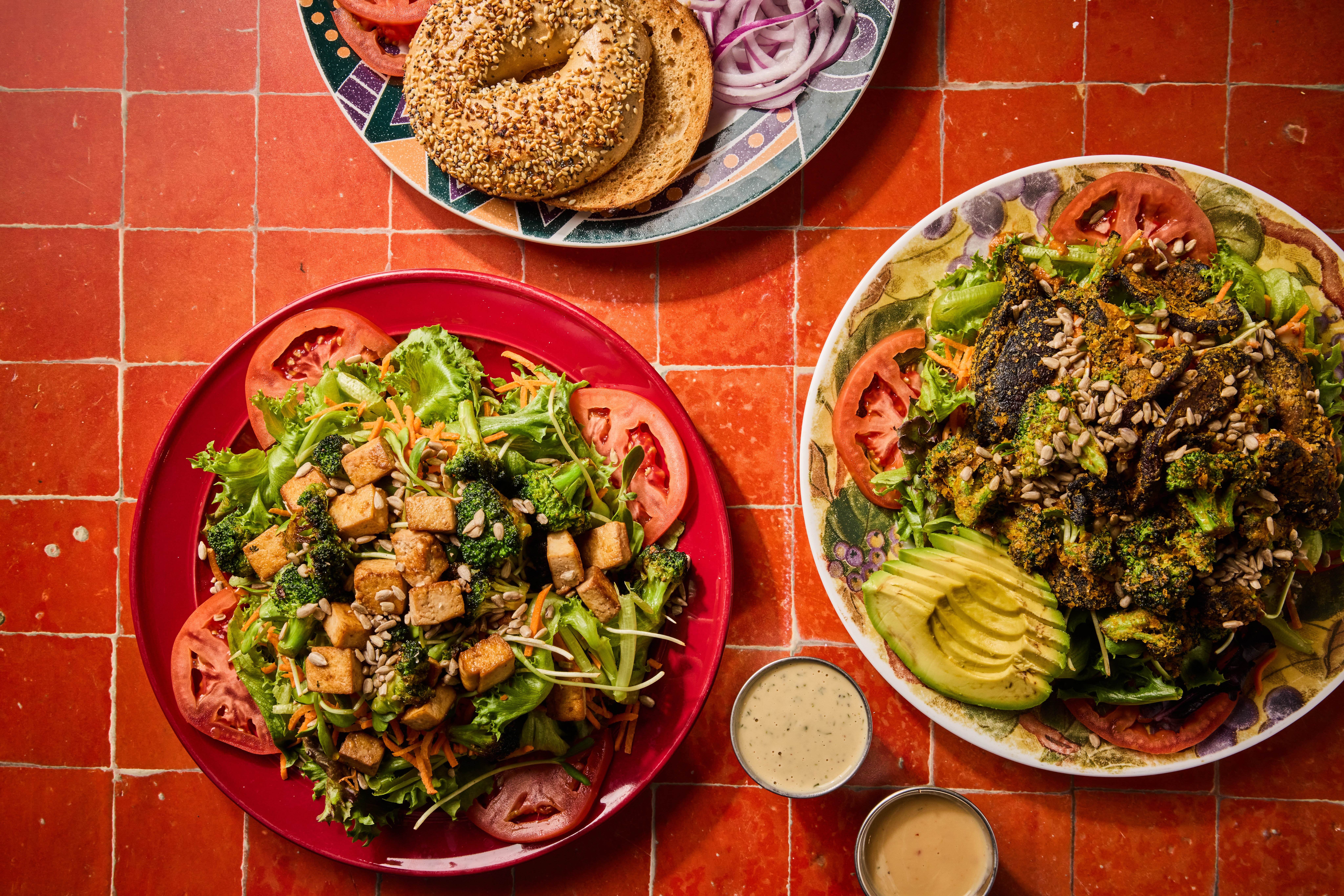 Two plates of salad with tomatoes, cucumbers, and greens, and a small cup of dressing on a red tiled table.