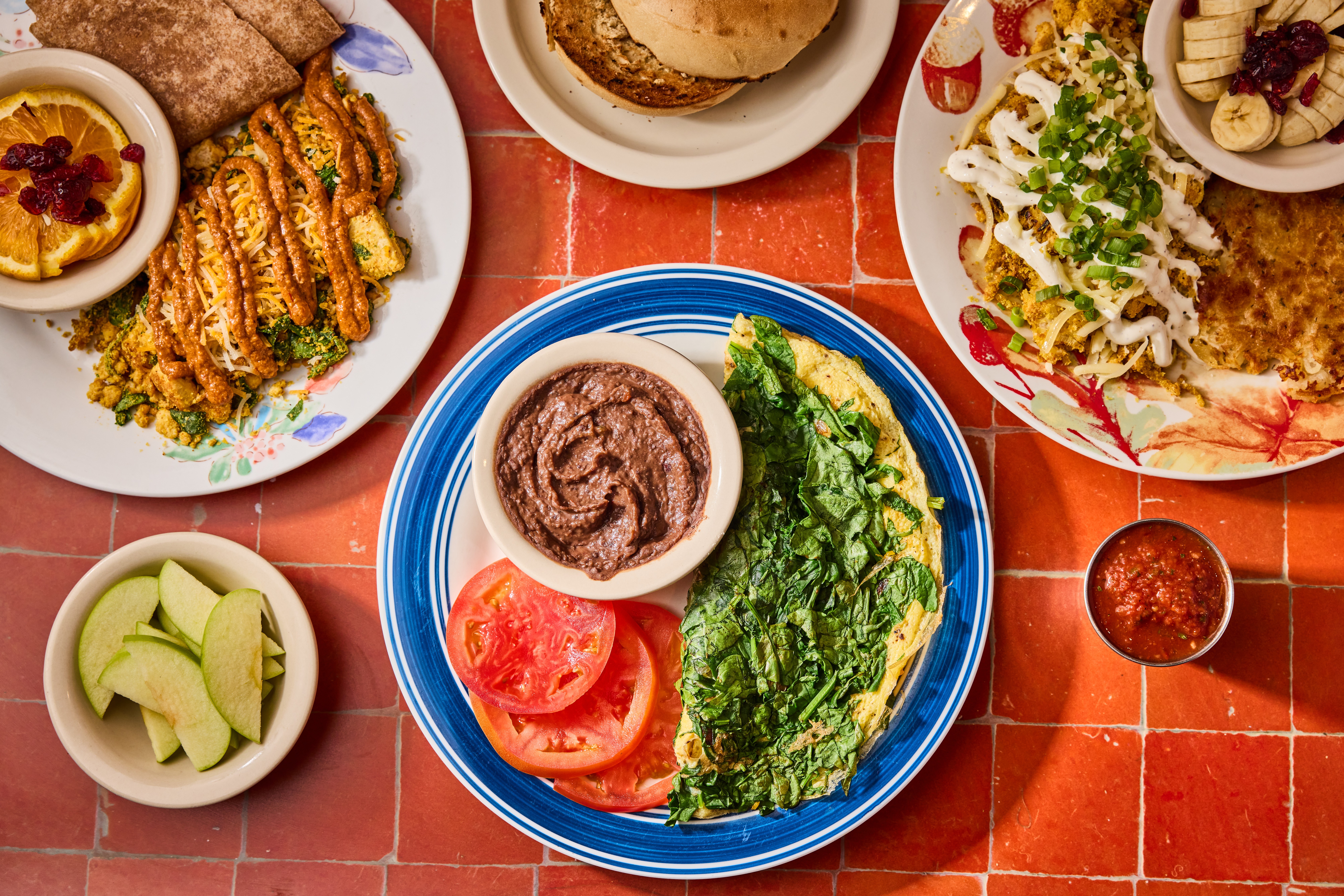 Top-down view of various dishes including fried food, salad, sliced tomatoes, and lime wedges on a red tiled table.