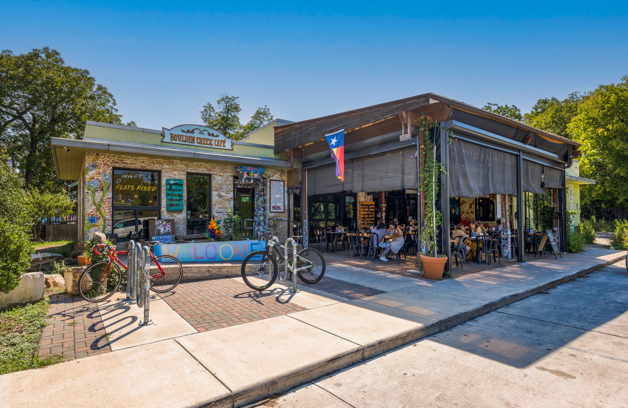 Two storefronts with outdoor seating and bicycles parked in front, trees and blue sky in background.