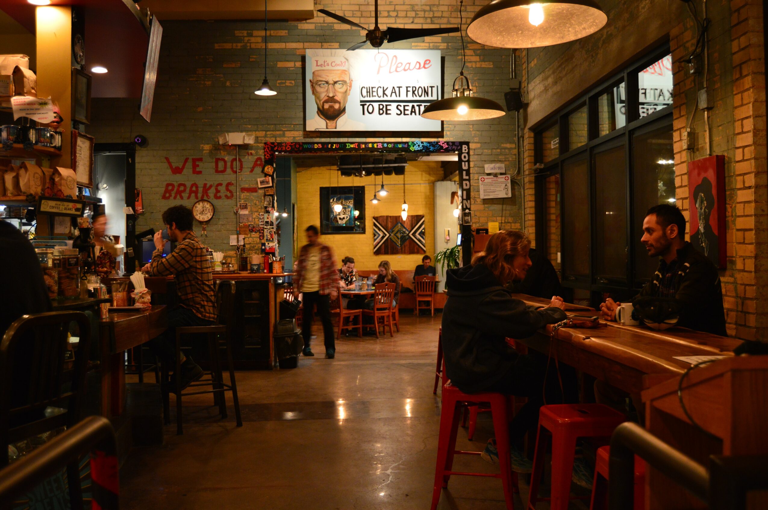 Interior of a cozy restaurant with people sitting at tables and a bar, warm lighting, brick walls, and a large window.
