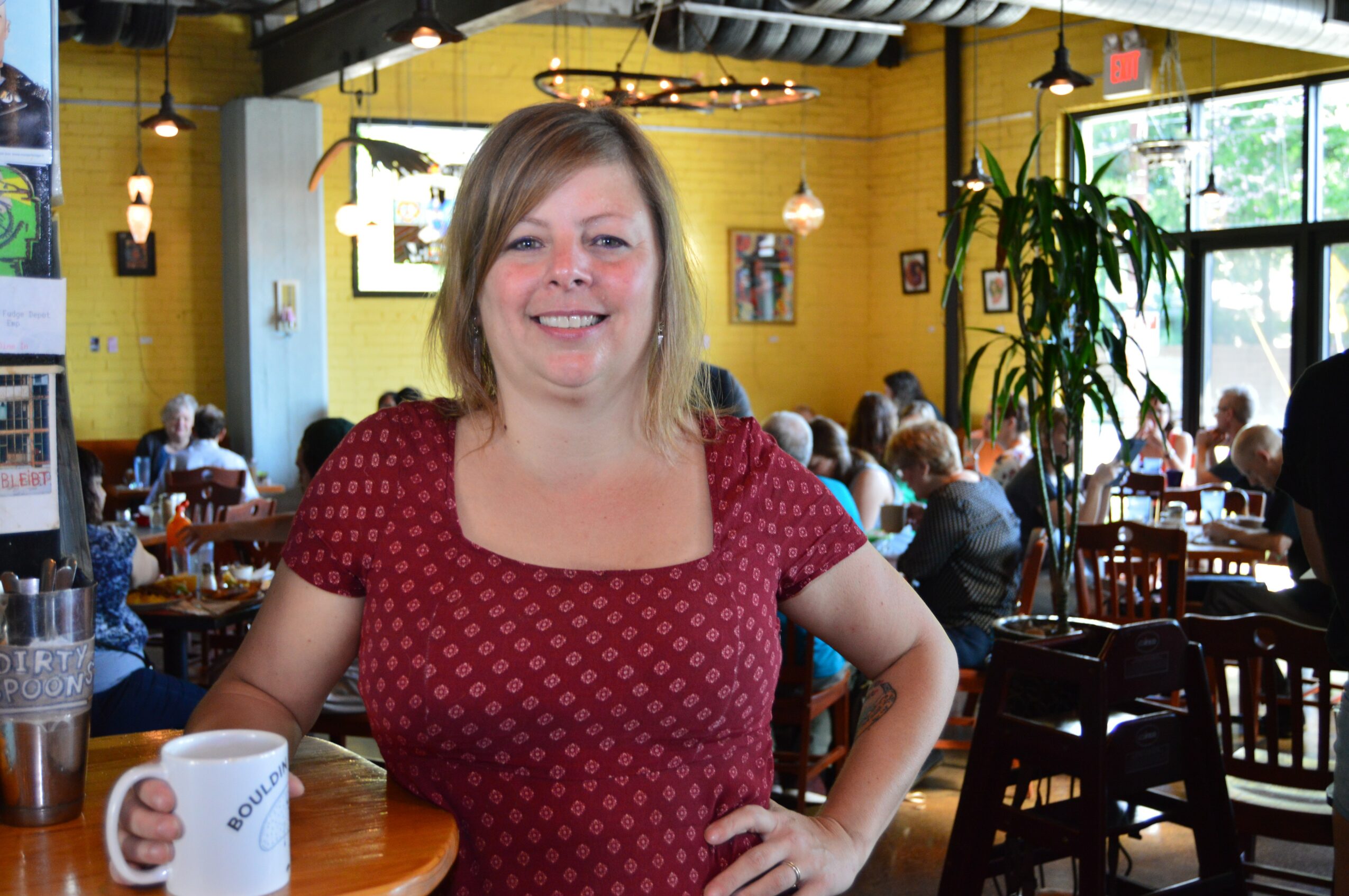 Smiling woman with shoulder-length hair in a red patterned top sitting at a restaurant table, with other diners and yellow walls in background.