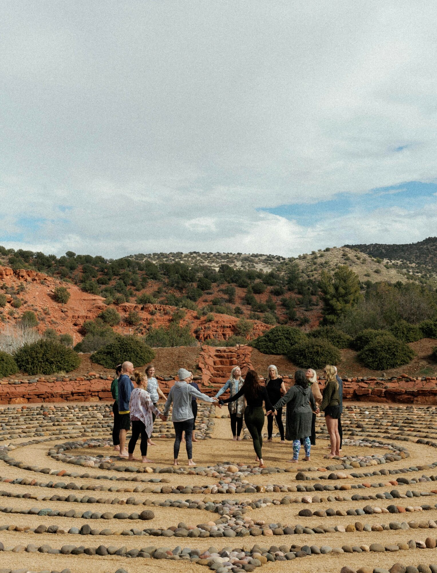 Group of people holding hands in a circle outdoors with red rocky hills and cloudy sky in background.