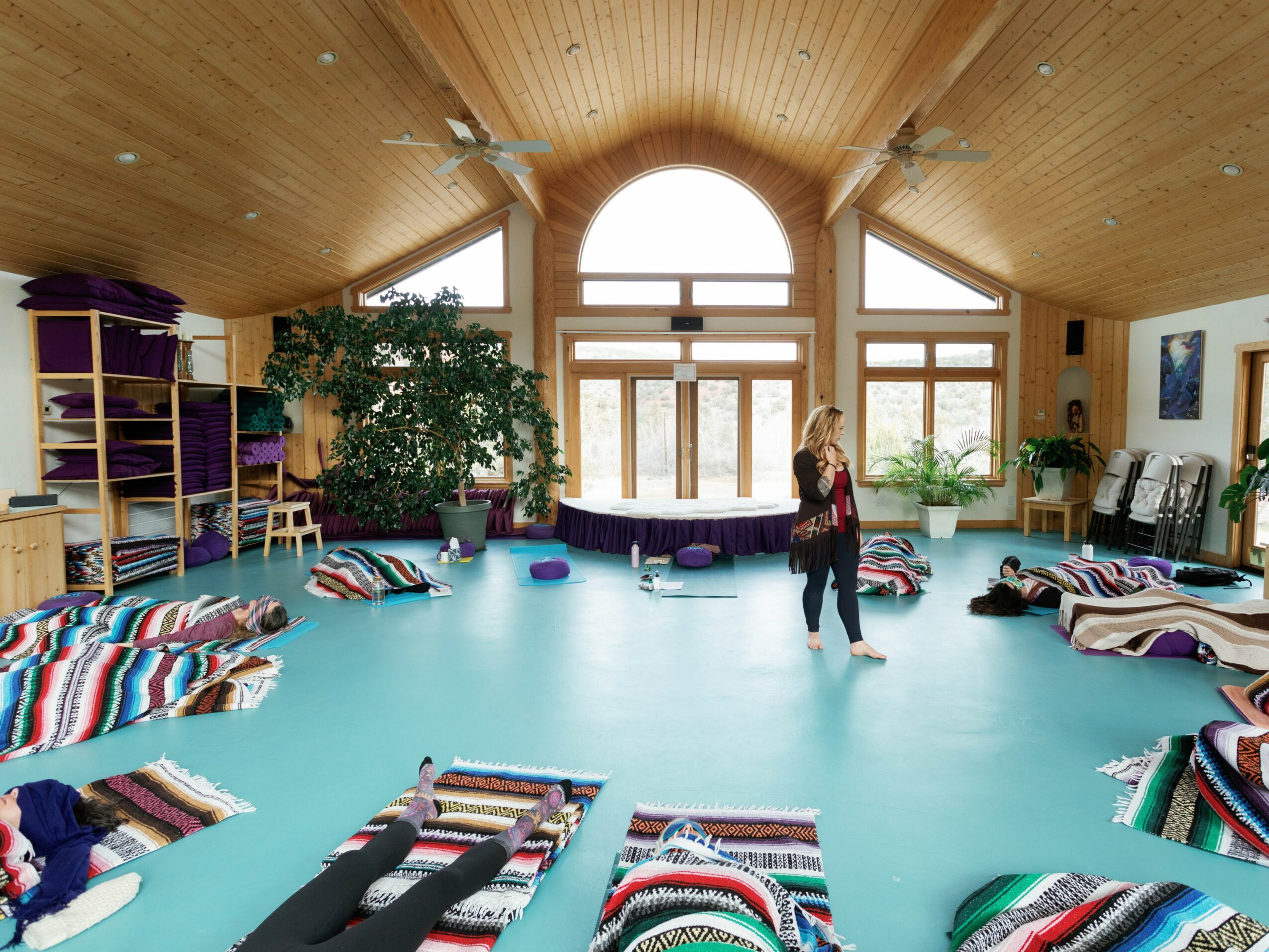 Spacious room with wooden ceiling, large window, colorful cushions on the floor, and a person standing near plants.