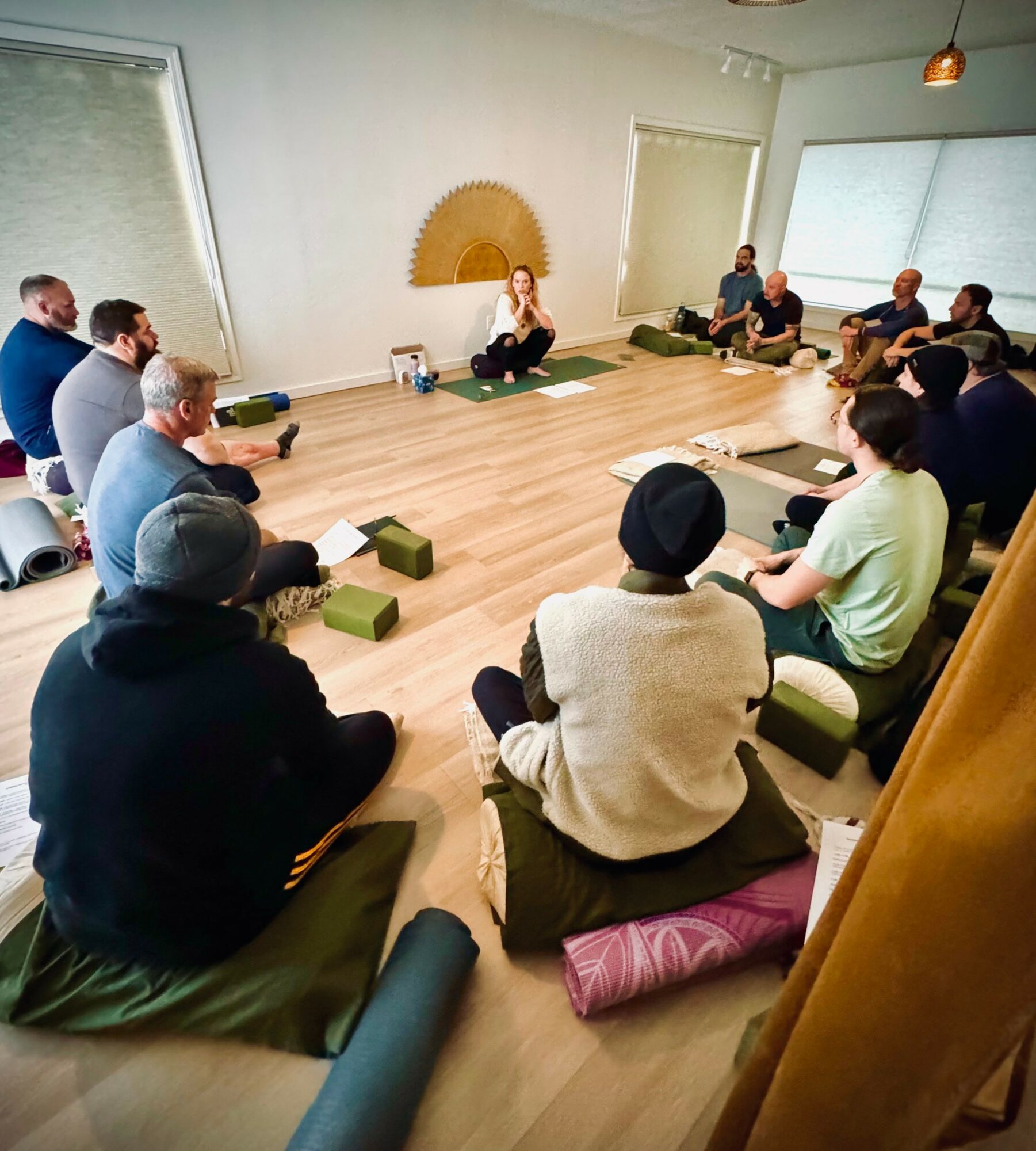 Group of people sitting on cushions in a circle in a bright room with large windows, listening to a woman speaking.