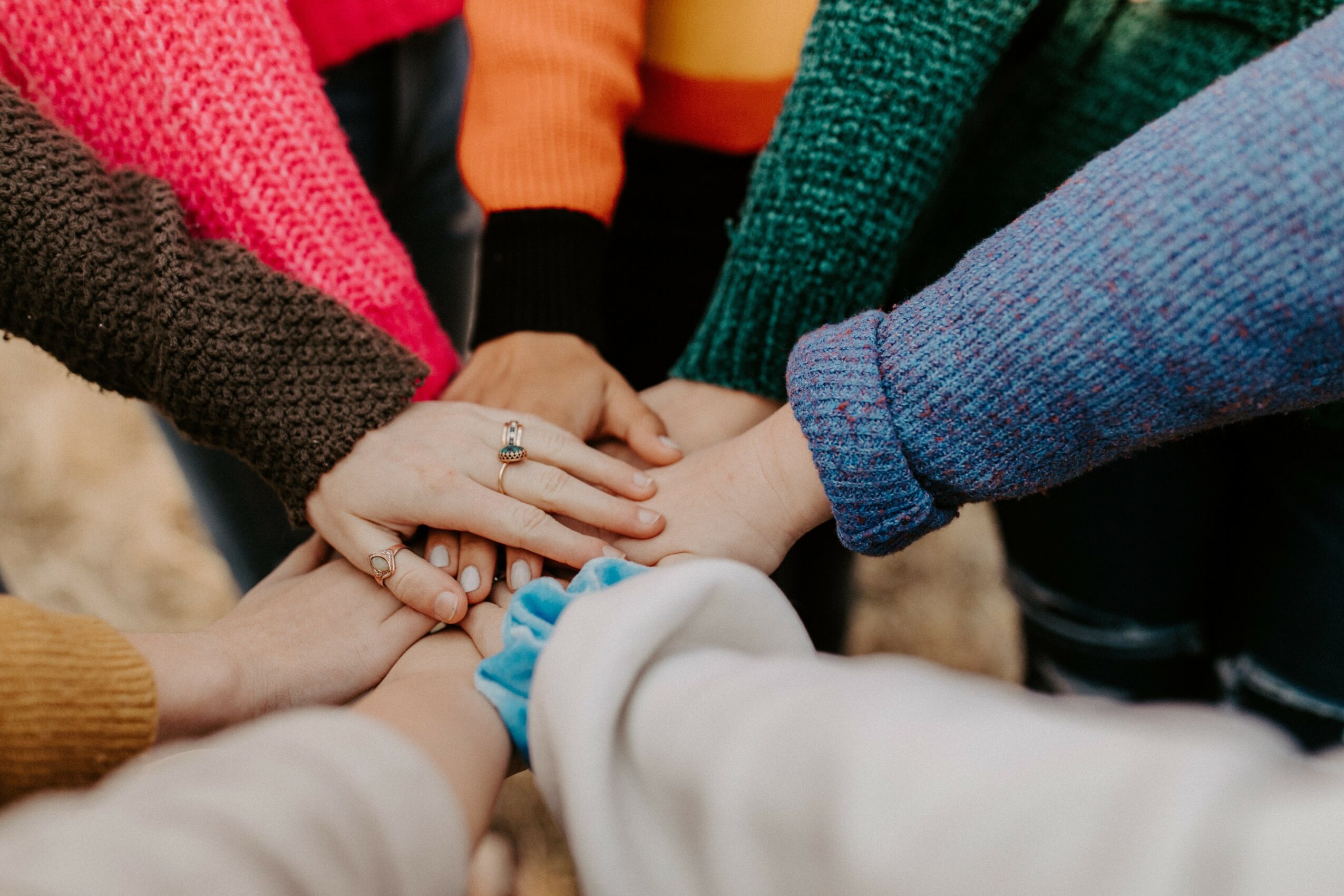 Multiple hands stacked together, showing unity and teamwork, with colorful clothing visible in the background.