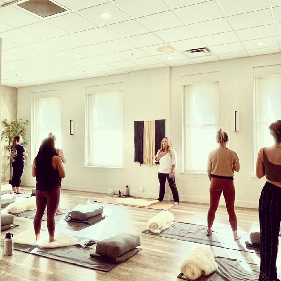 Group of people participating in a yoga or meditation class in a bright, spacious room with large windows and mats on the floor.