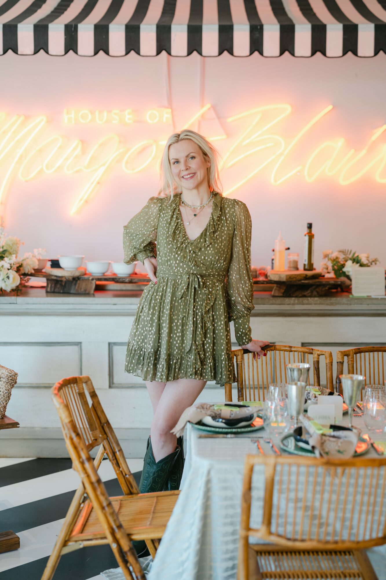 Young woman in a green dress standing at a restaurant table with a neon sign background, smiling, with table settings and chairs visible.