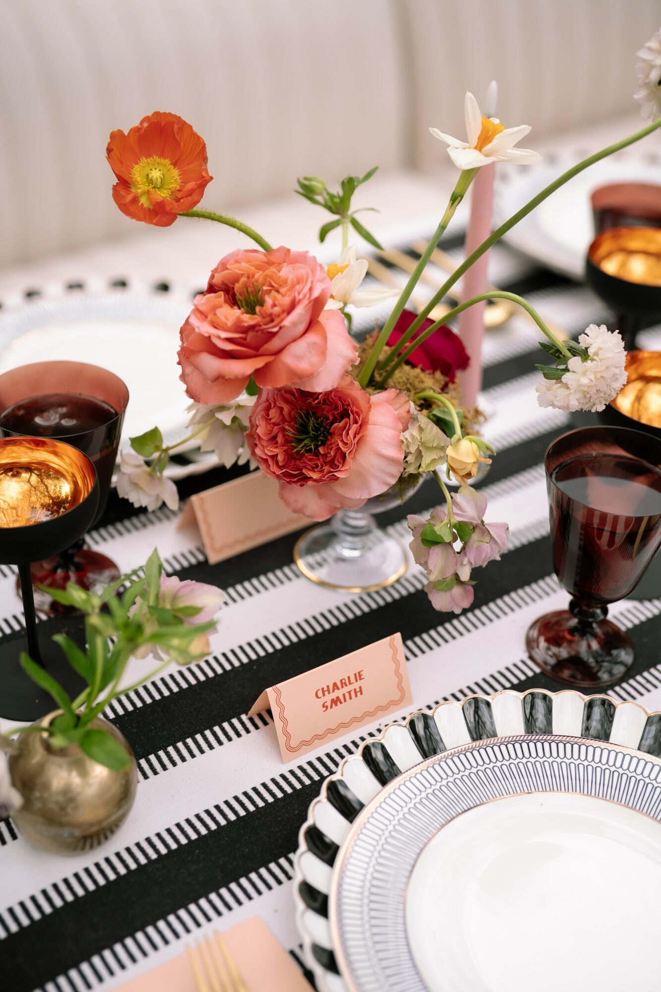 Floral centerpiece with pink, white, and orange flowers on a black and white striped tablecloth, with glassware and place cards.