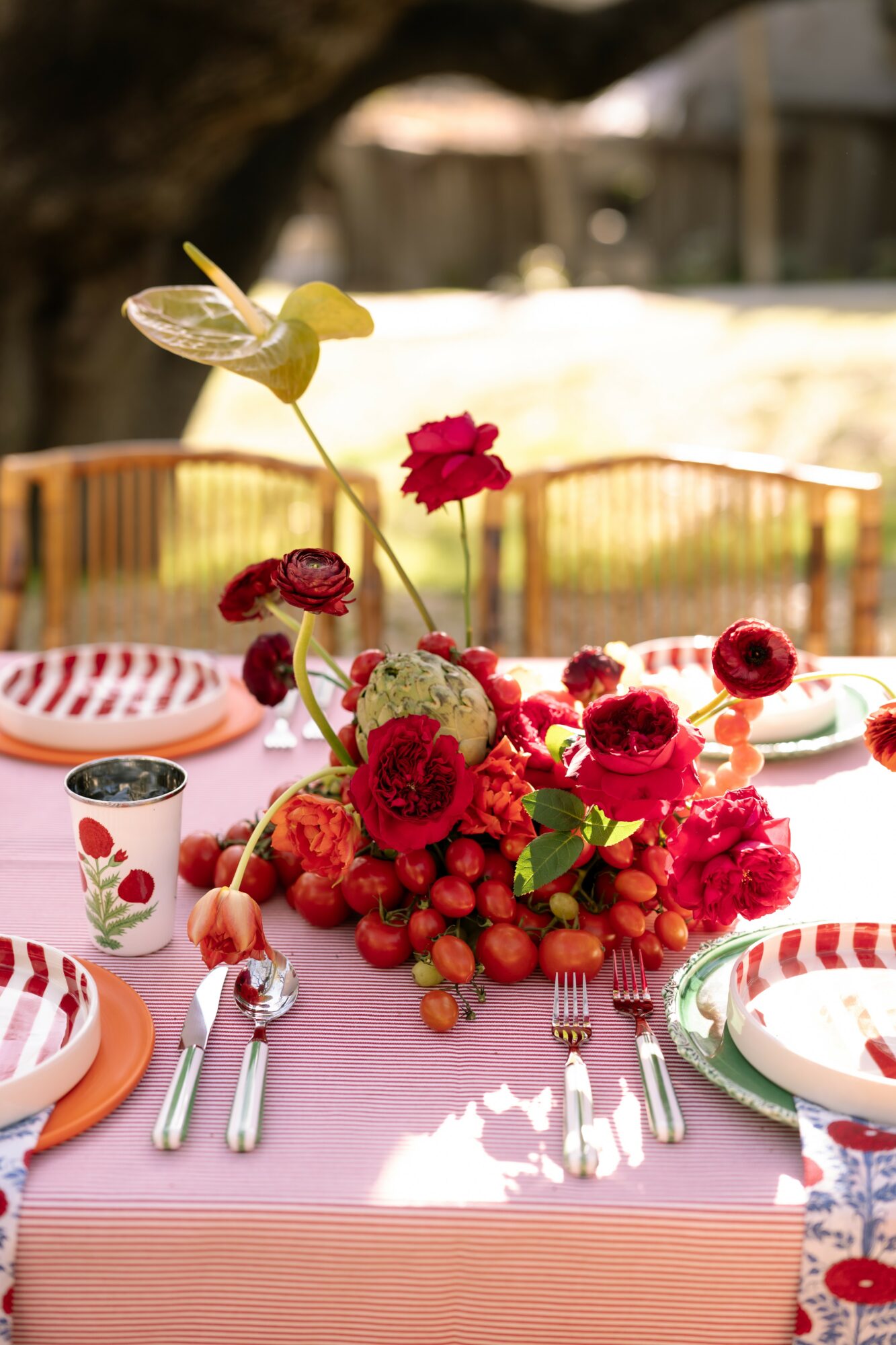 Table set outdoors with a pink tablecloth, floral centerpiece, plates, and cutlery, with a blurred background of trees and structures.