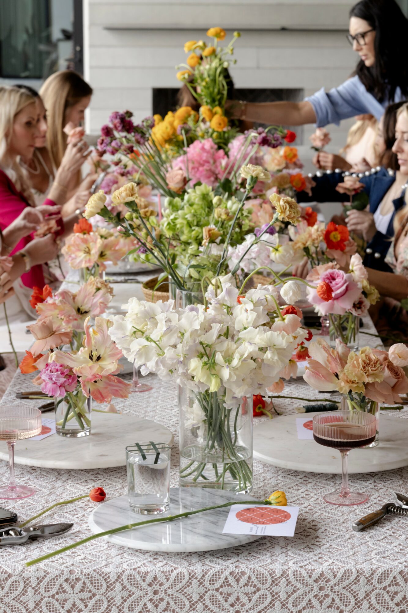 People gather around a table with a large floral centerpiece, flowers in various colors, and tableware, outdoors.