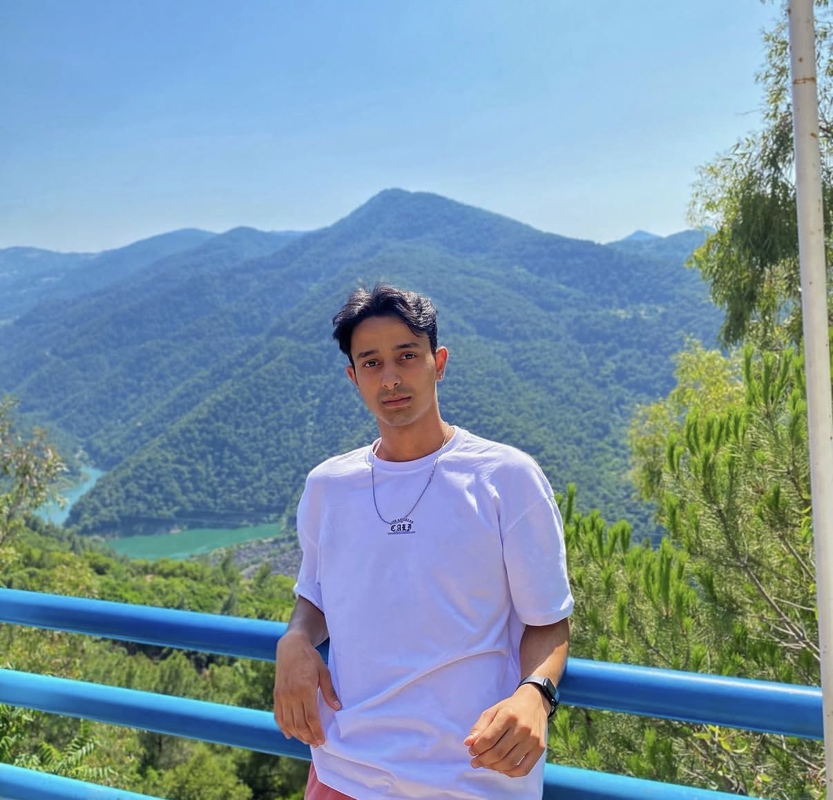 Young man standing outdoors with mountains and trees in background, blue sky, wearing a white t-shirt and necklace.