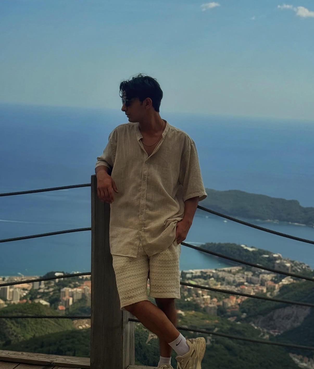Young man with dark hair in beige shirt and shorts leaning on a railing overlooking a coastal landscape with water and land.