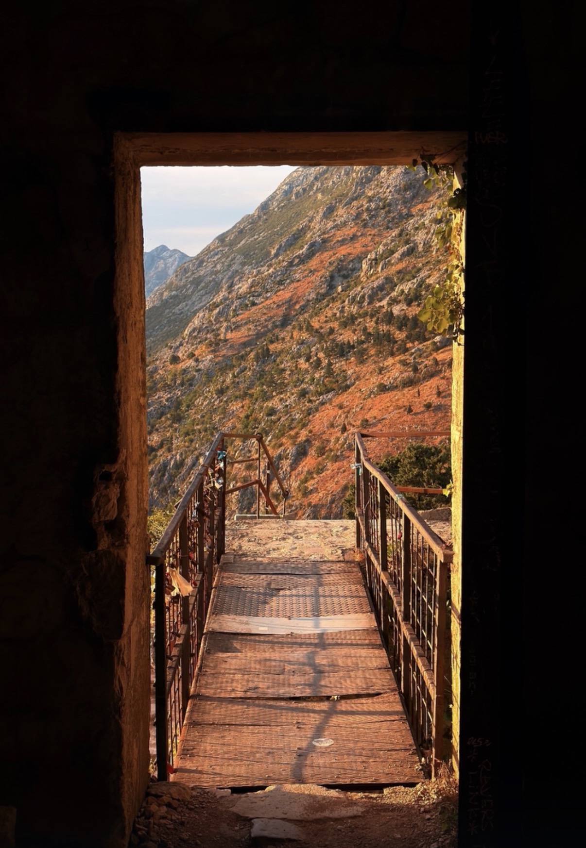 View through a doorway showing a wooden bridge with metal railings over a mountain landscape with autumn foliage.