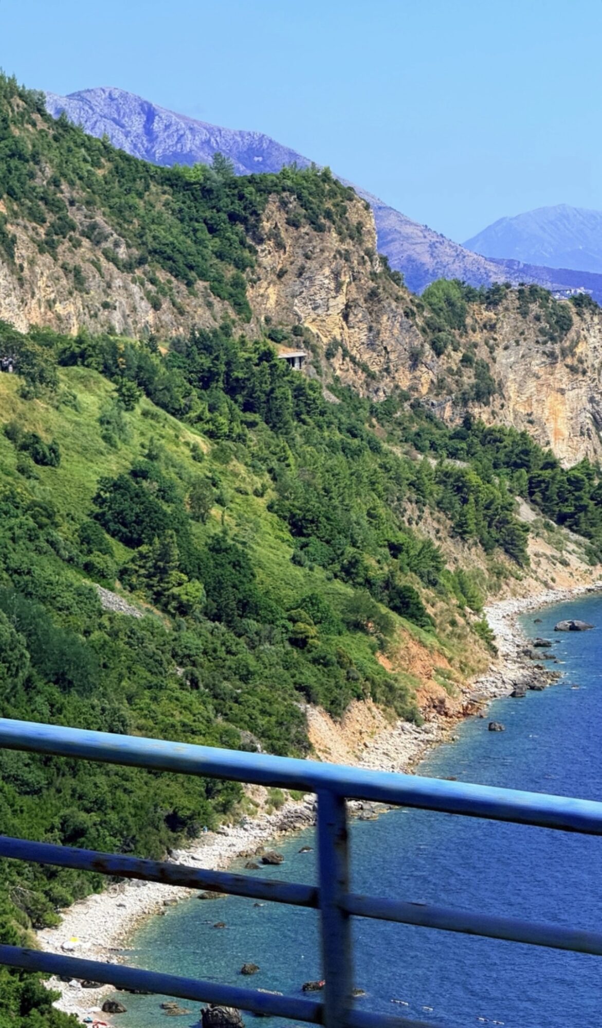 Coastal landscape with green hills, rocky cliffs, and blue water, viewed from a railing on a bridge or walkway.