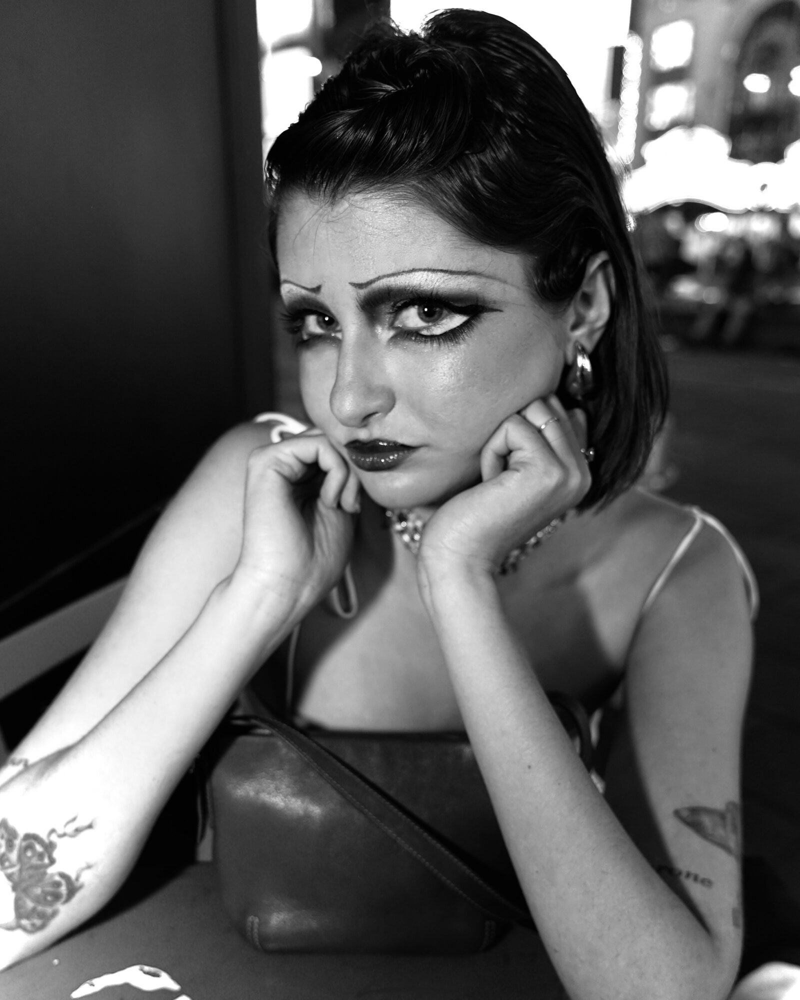 Woman with dark hair and makeup resting her face on her hands, sitting indoors with a blurred city background.