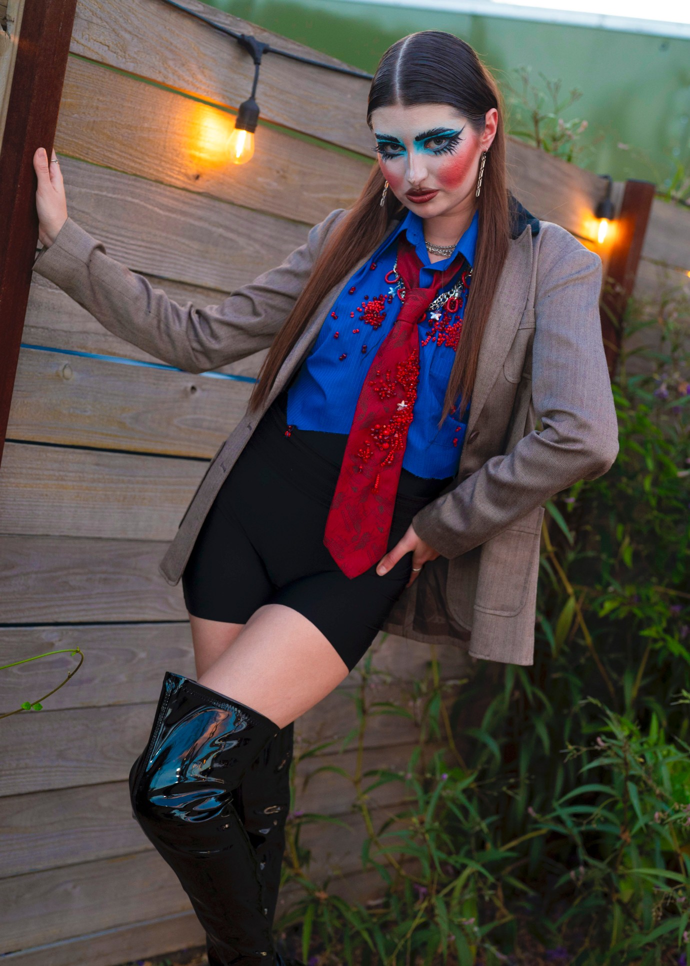 Person with dramatic makeup and long hair, wearing a blazer, blue shirt, red scarf, black shorts, and thigh-high boots, outdoors at night.