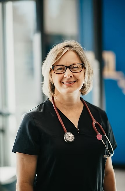 A smiling woman with glasses and short blonde hair wearing a black medical uniform and stethoscope, standing indoors.