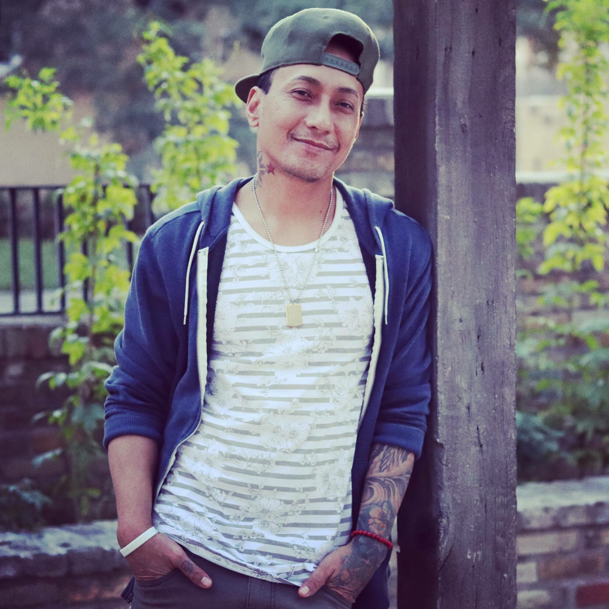 Young man outdoors wearing a cap, striped shirt, and blue hoodie, smiling, with trees and a wooden post in background.
