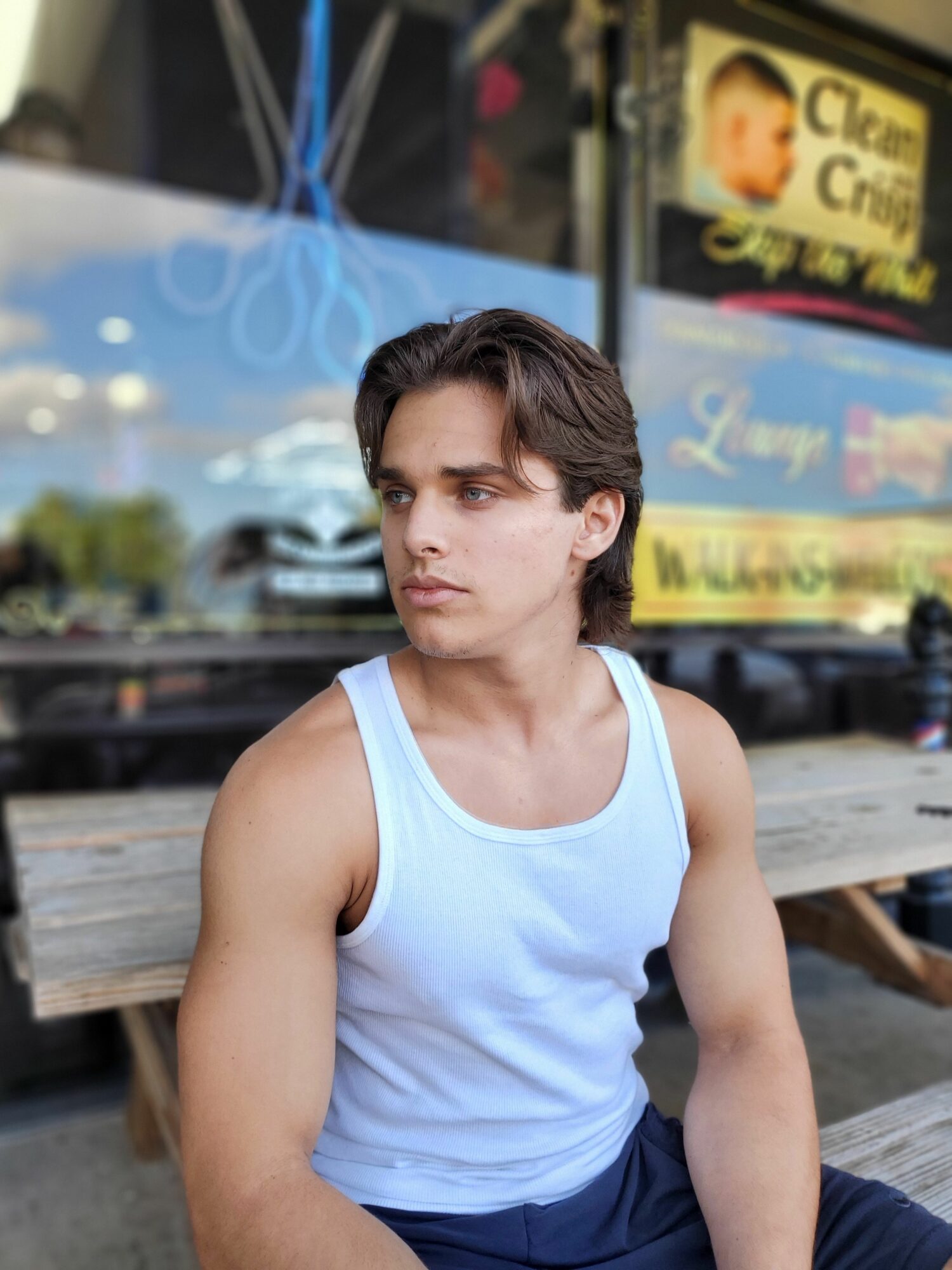 Woman with short dark hair in a white sleeveless top sitting on a bench outdoors, looking down, with a blurred city background.