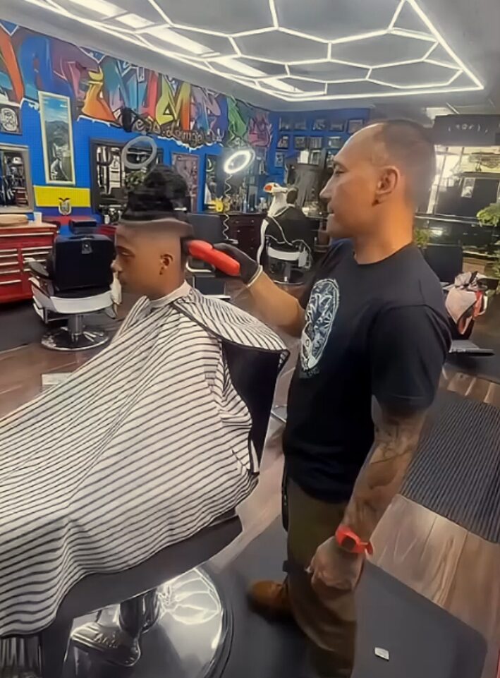 Barber trims a young boy's hair in a colorful, modern barbershop with geometric ceiling lights and vibrant wall art.