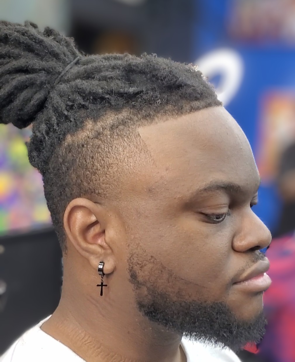 Side profile of a man with dreadlocks and a beard, wearing a cross earring, in a public setting.