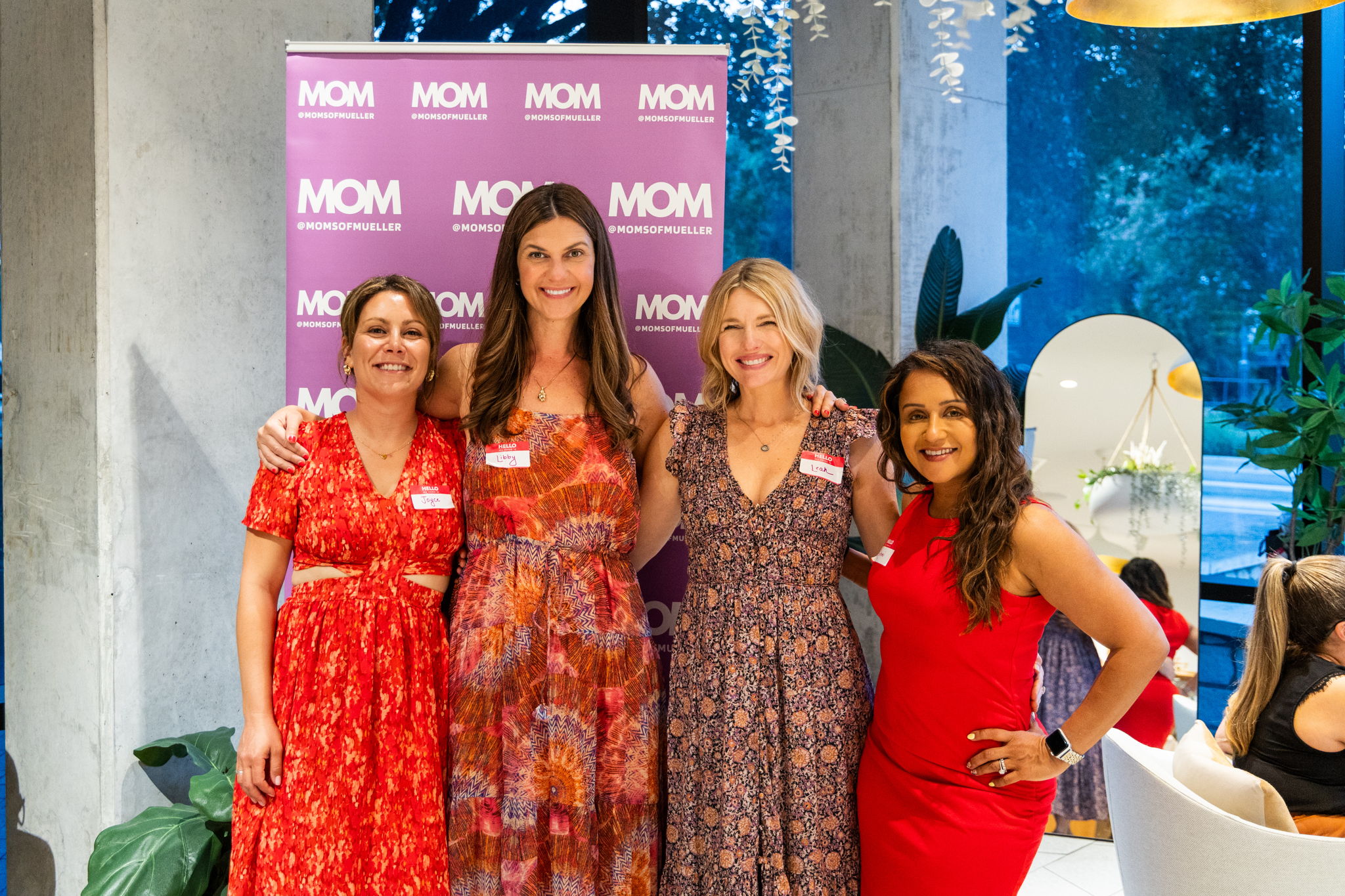 Four women standing together indoors, smiling, with a pink MOM banner in the background, and a large window with blue sky outside.