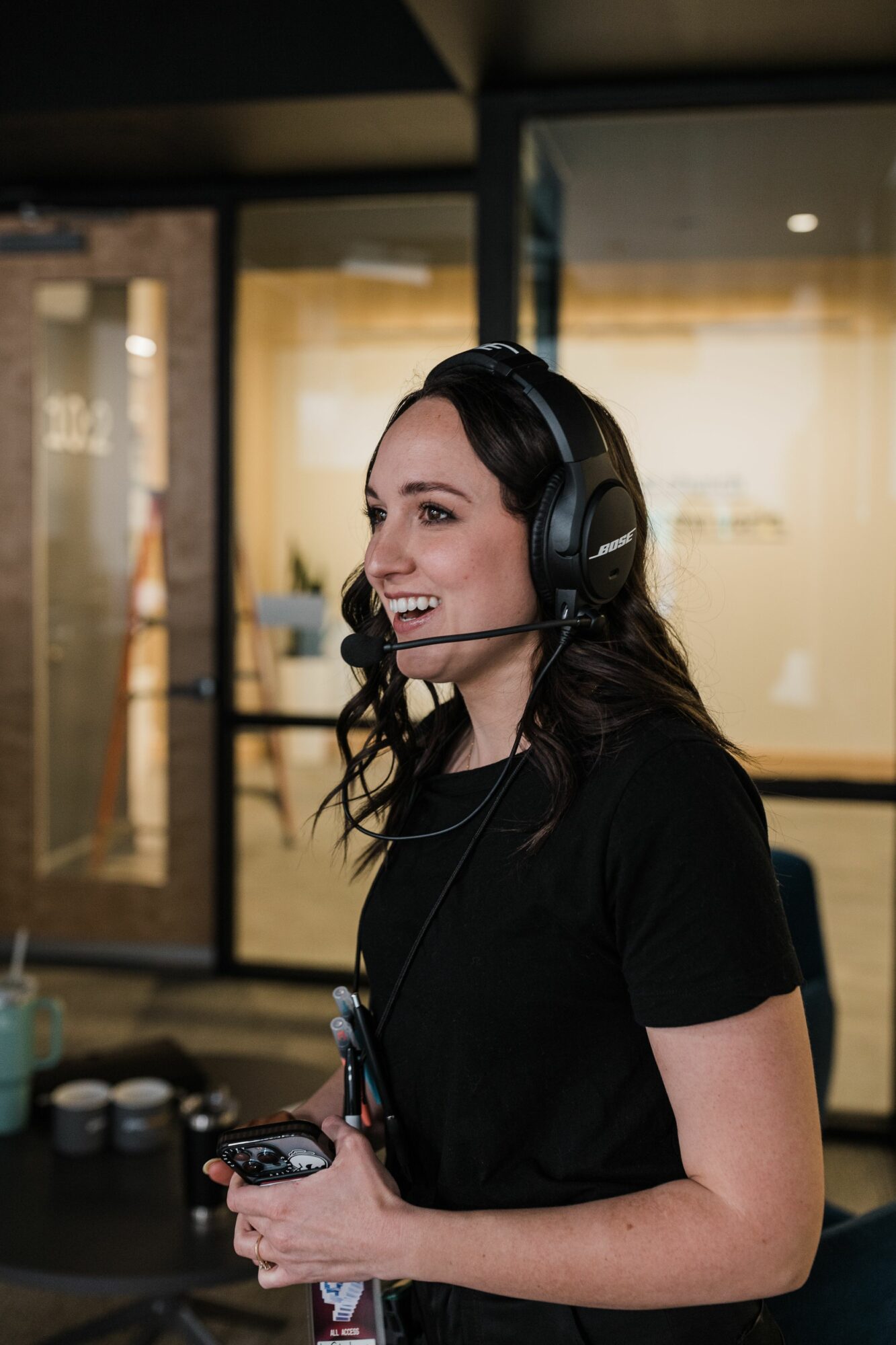 Woman wearing a headset, smiling, holding a device, in an indoor setting with glass doors behind her.