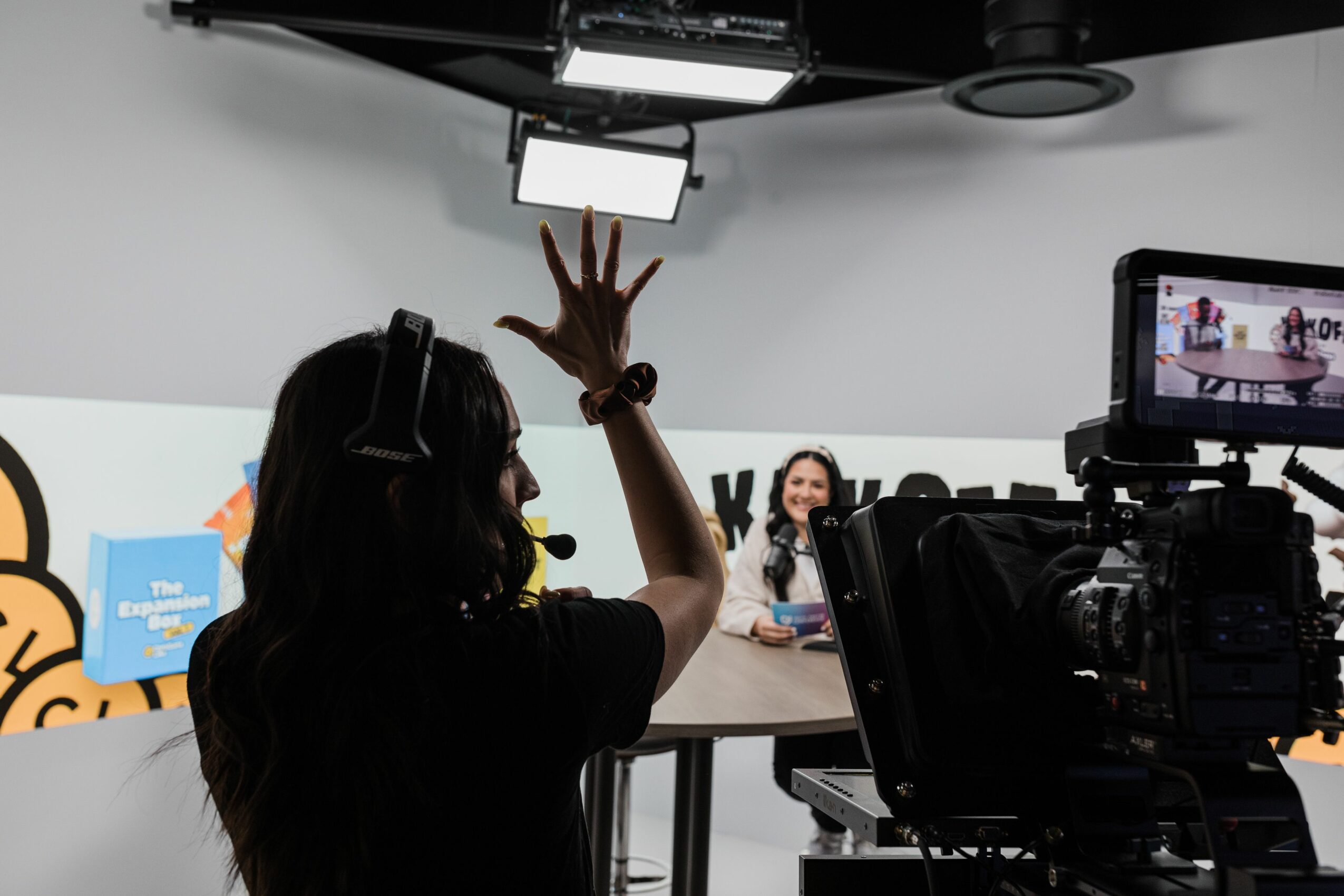 Person with long hair and headphones raising hand in a studio with cameras and a monitor, two women sitting at a table.