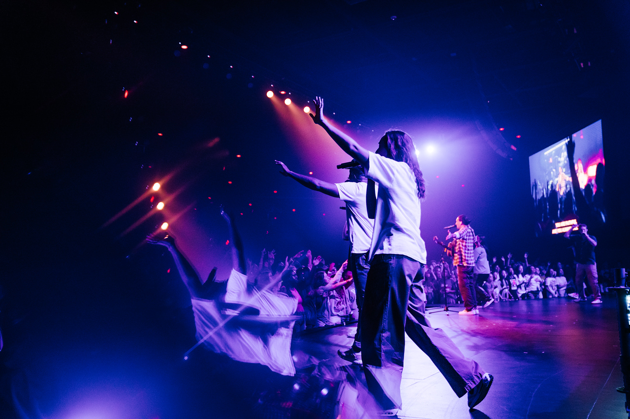 People on stage with one person reaching out, colorful lights, and a large screen in the background, concert or event setting.