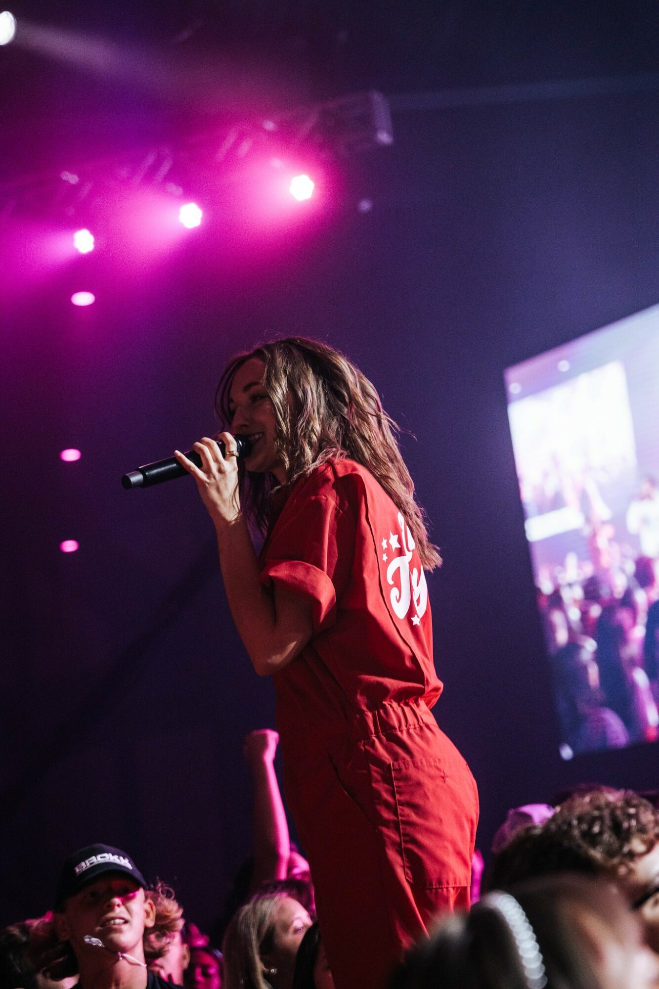 Person singing into a microphone on stage with pink and purple lighting, wearing a red outfit, audience visible in background.