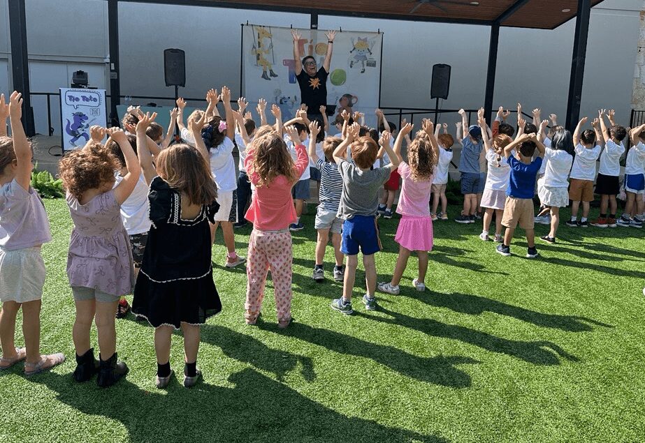 Children participate in outdoor activity with a person on stage, raising hands, on a grassy area under a roofed structure.