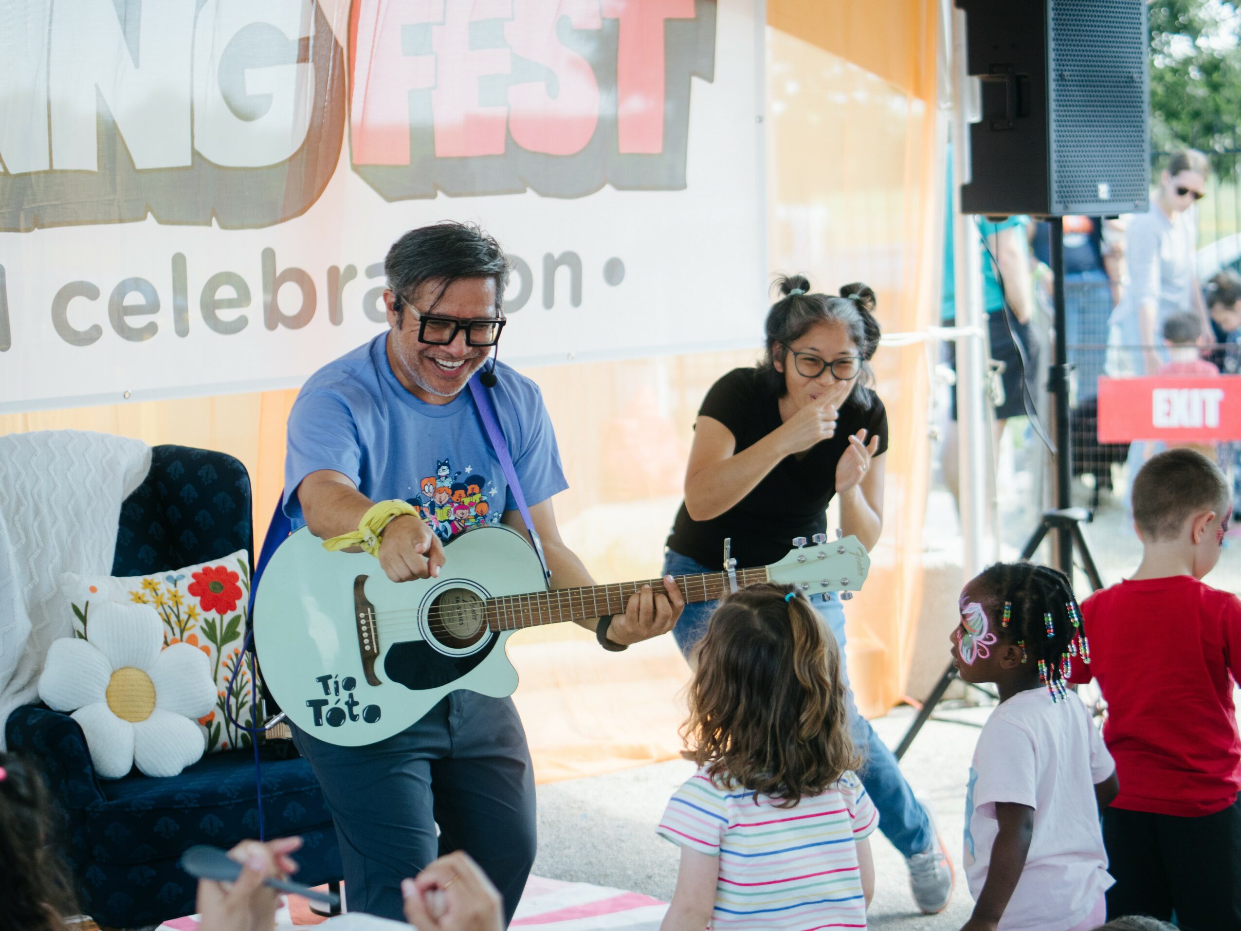 Two women perform on stage with children watching, one playing guitar and the other singing, outdoors during daytime.