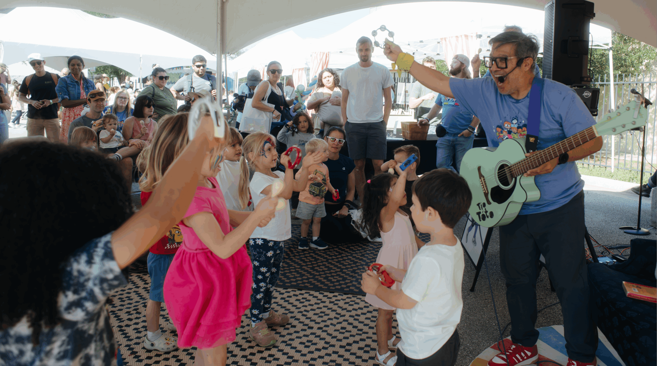 Man with sunglasses plays guitar and sings to children dancing and clapping outdoors under a tent, with a crowd watching.