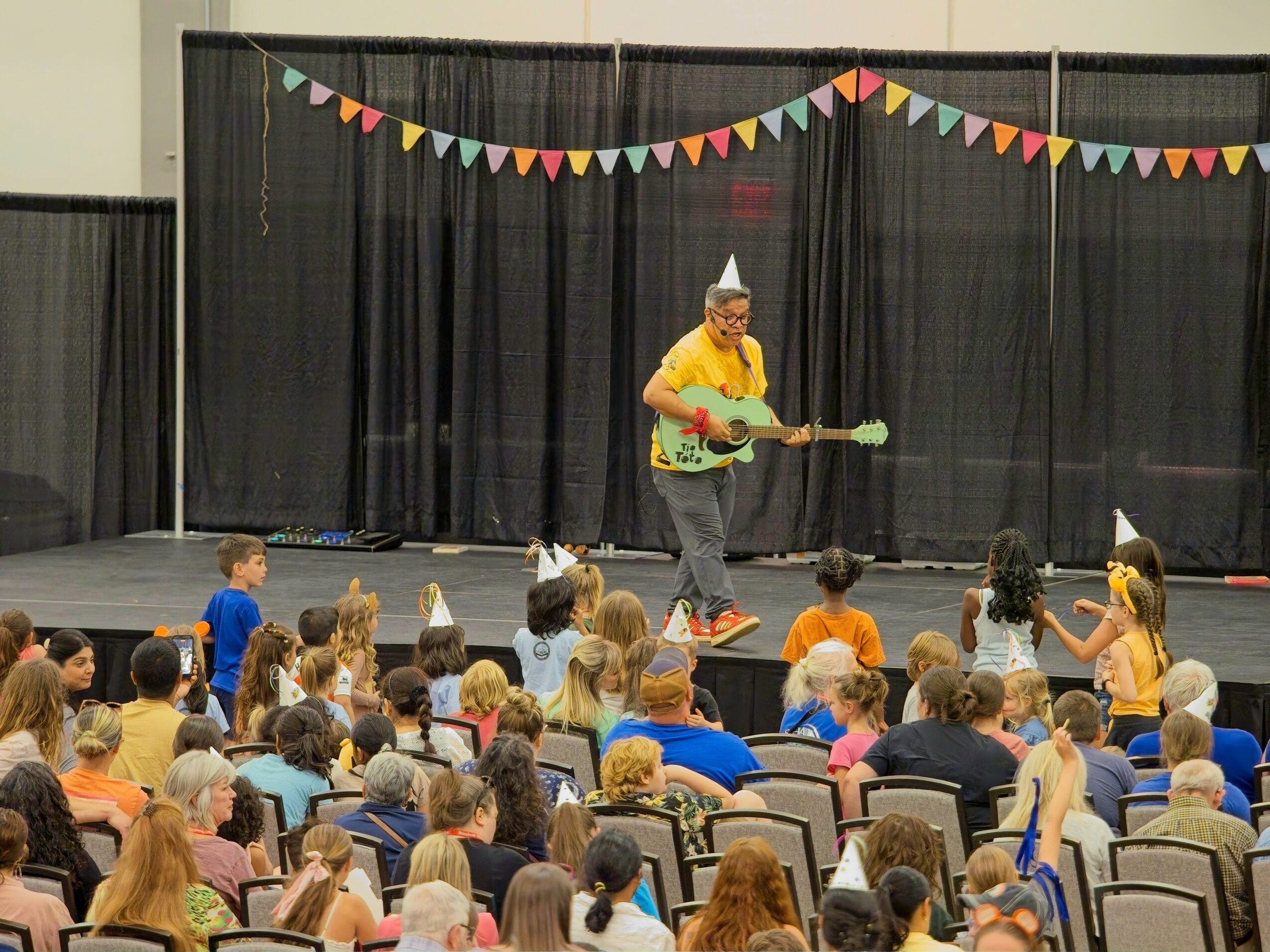 Person wearing a party hat plays guitar on stage with children watching, black curtains and colorful bunting in background.