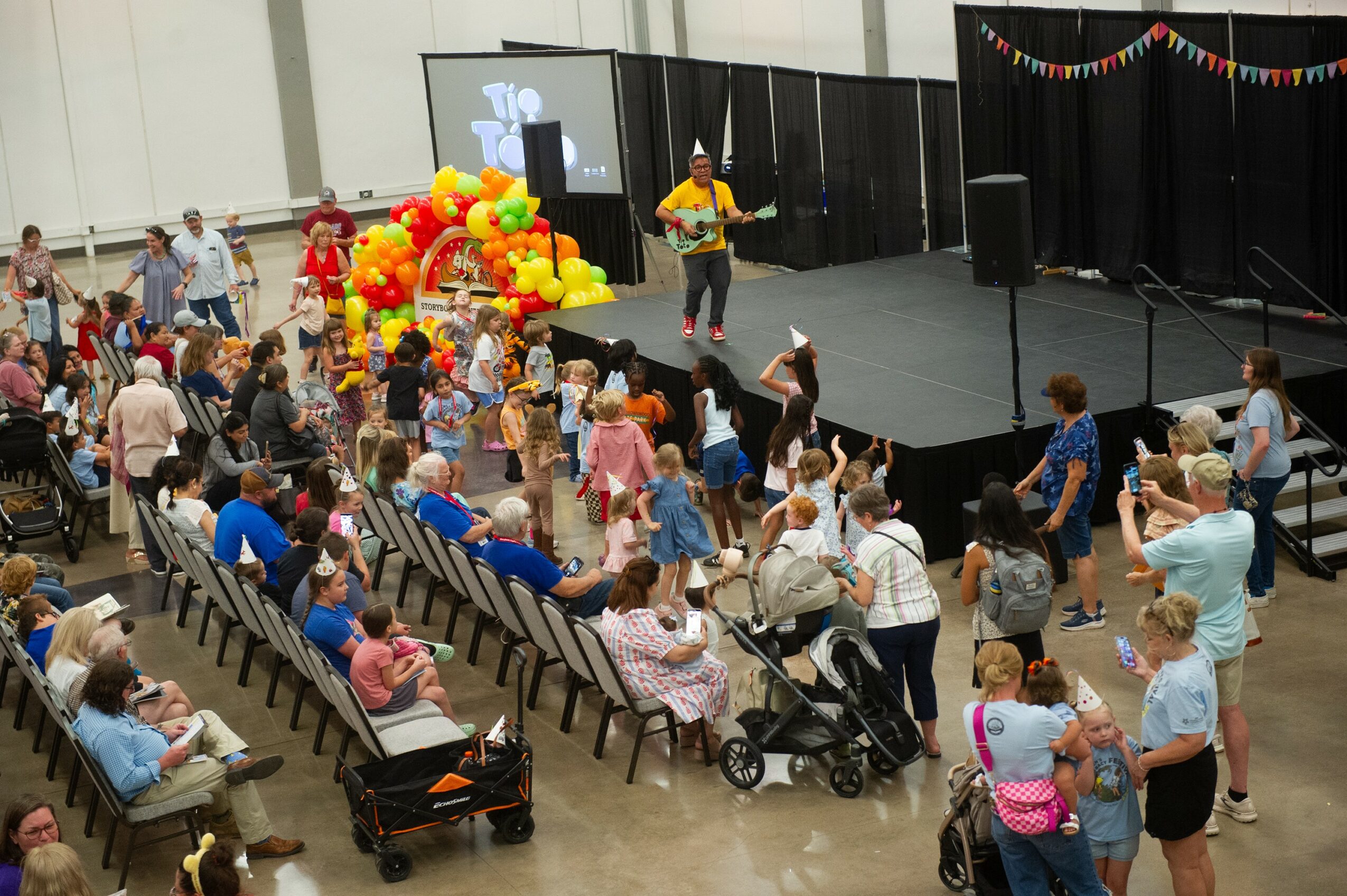 Crowd of children and adults watching a performer on stage decorated with balloons at an indoor event.