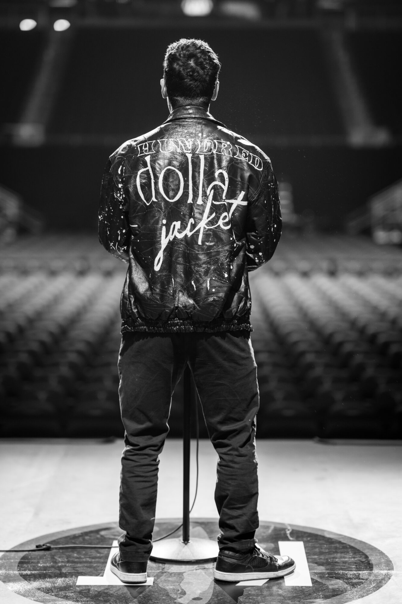 Person standing on stage with back facing camera, wearing a jacket with text, in an empty auditorium.