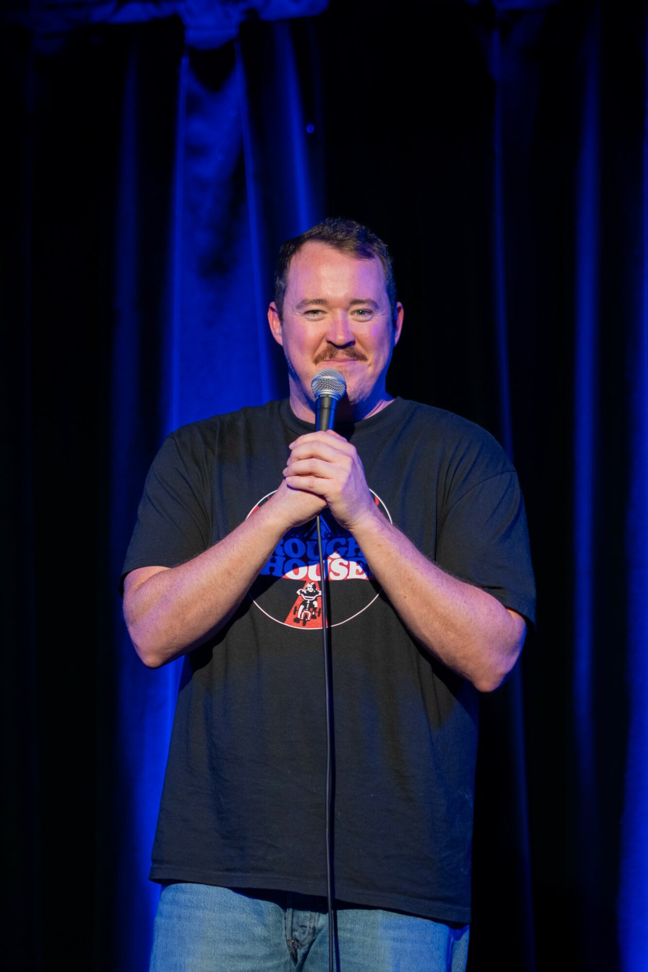 Man holding microphone on stage with dark curtains behind him, smiling, wearing a black T-shirt and jeans.