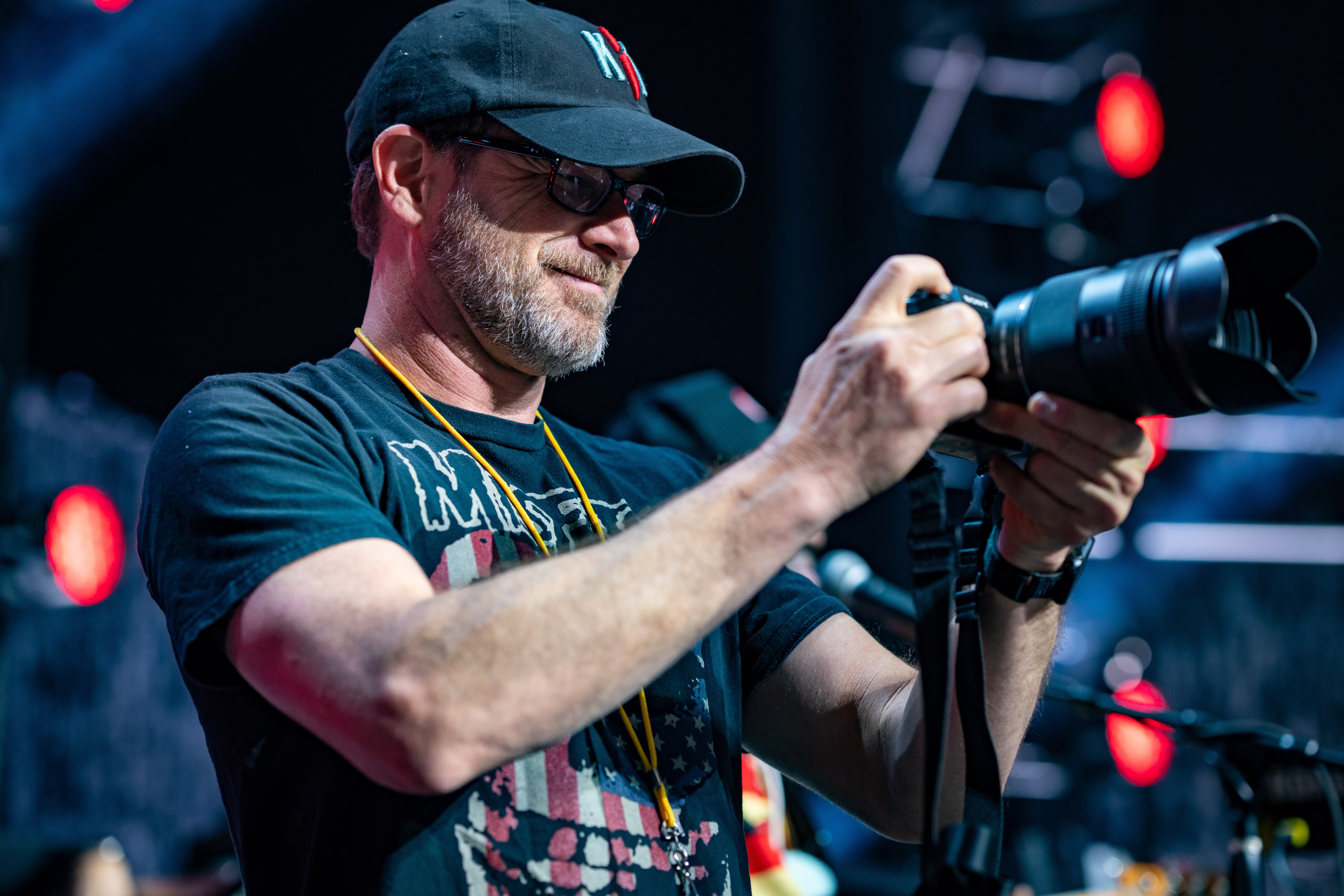 Man with glasses and a cap holding a camera, focused on taking a photo at an event, with stage lights in background.