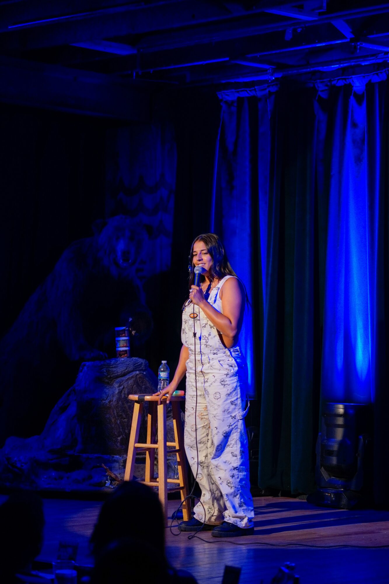 Person standing on stage with a microphone, wearing a patterned outfit, in front of blue curtains.