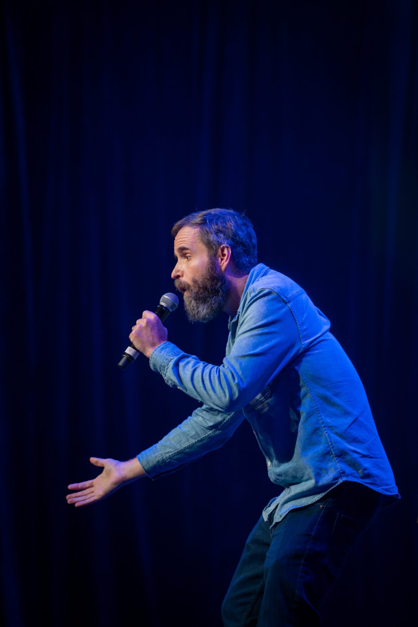 Man with a beard holding a microphone, gesturing with his other hand on stage with dark background.